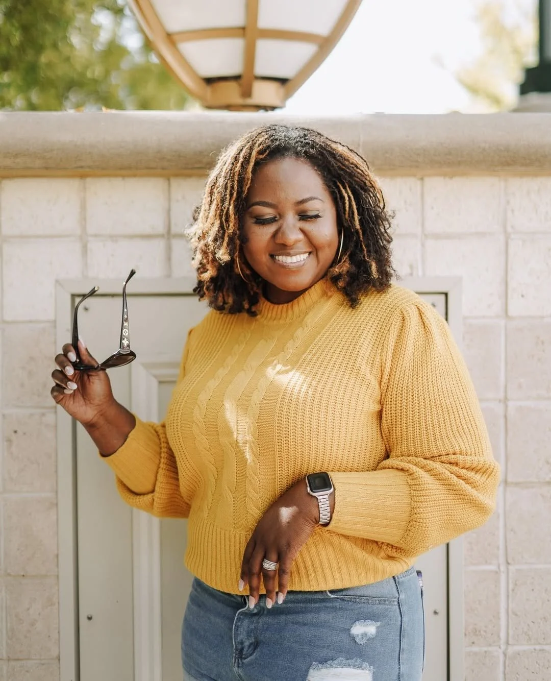 A woman with curly hair wearing a yellow sweater and ripped jeans, smiling and holding sunglasses, standing outdoors in front of a brick wall and a lamp. she is wearing a watch and a ring.
