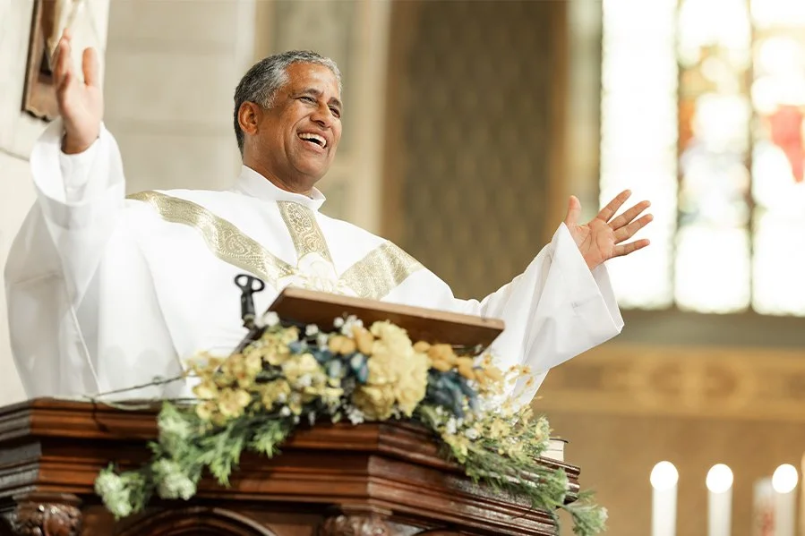 A priest wearing white robes with gold accents, smiling and raising his hands, standing behind a wooden pulpit decorated with flowers, inside a church.