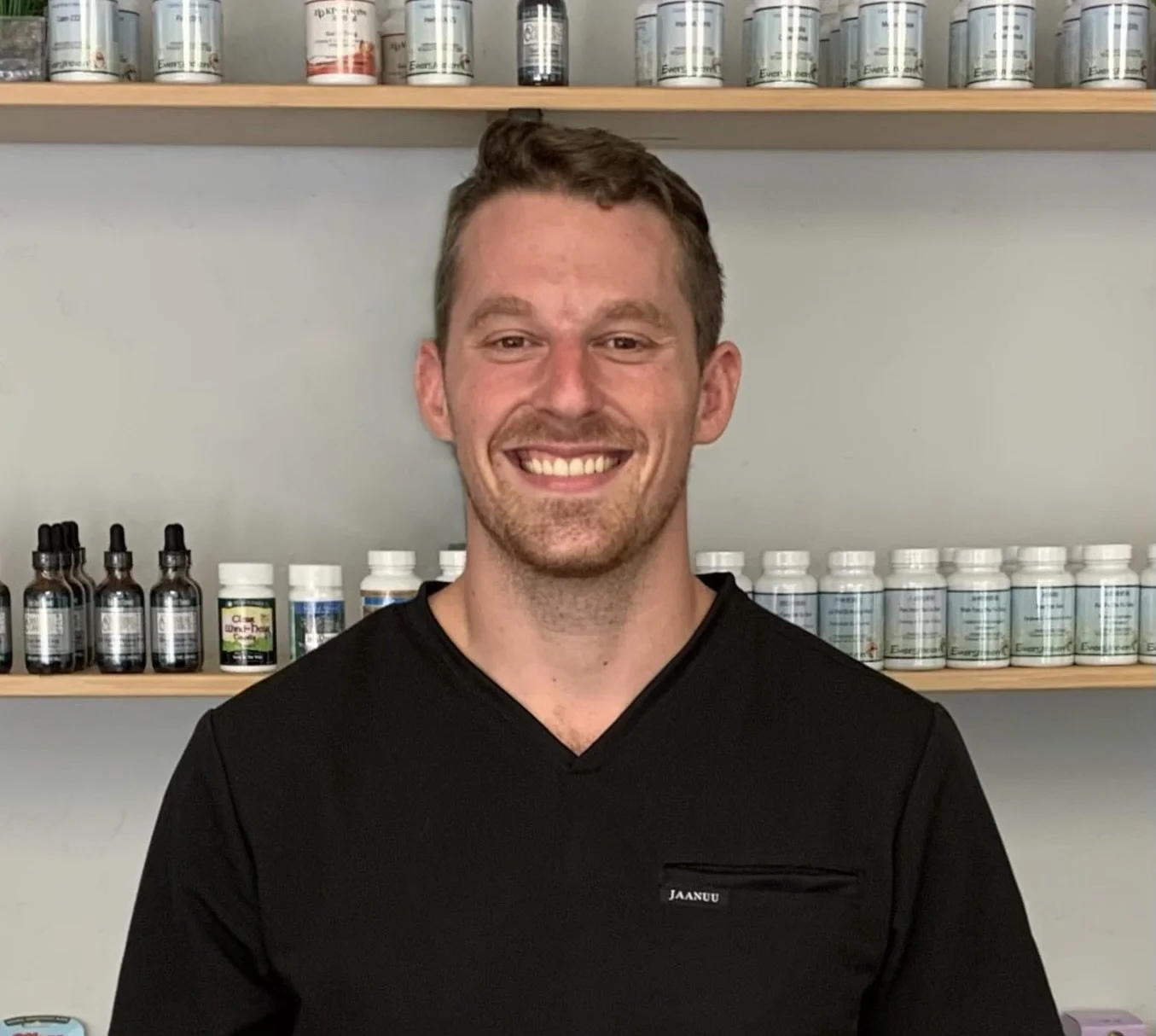 A smiling man with short brown hair and a beard, wearing a black shirt, standing in front of shelves filled with bottles and containers in a wellness or supplement store.