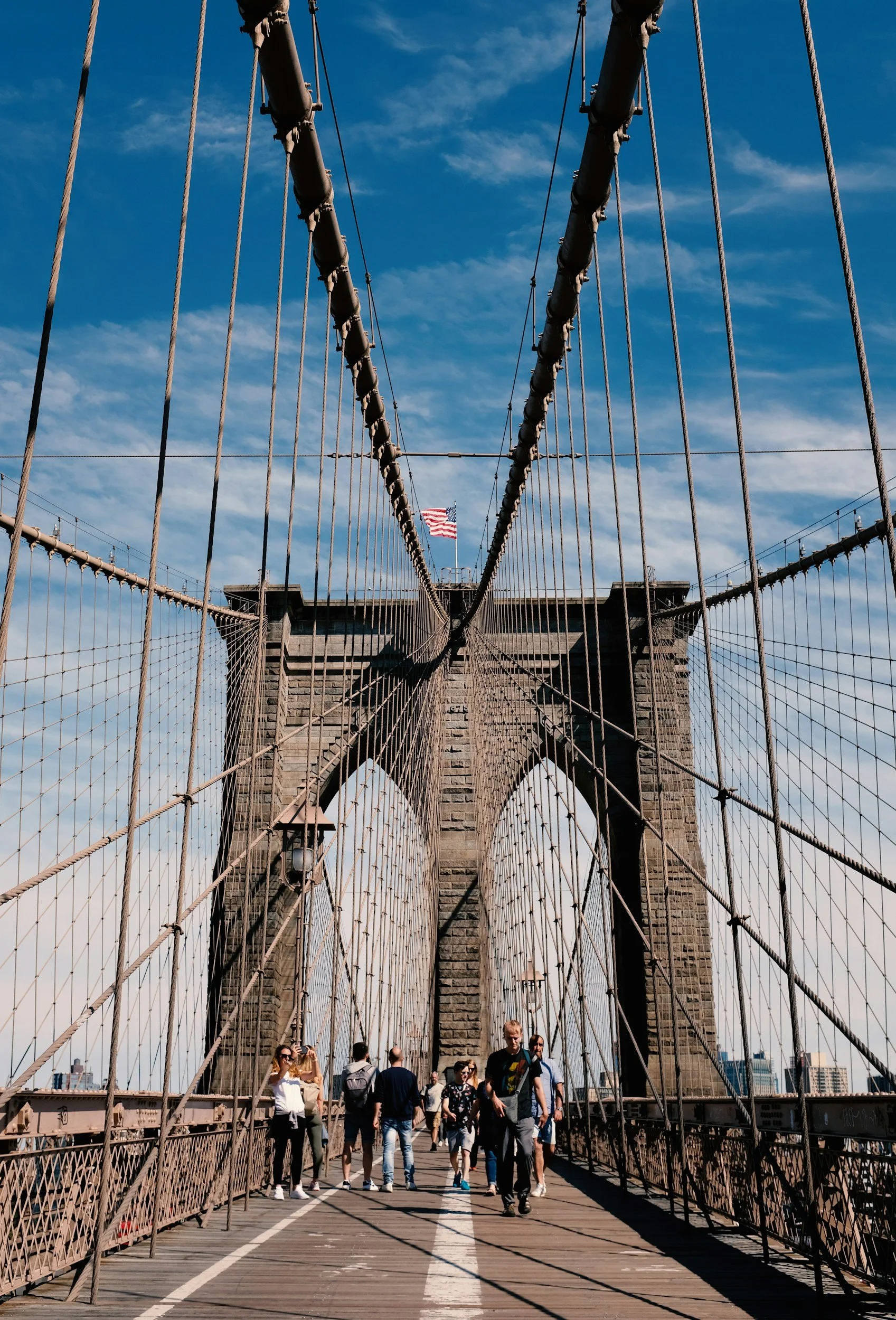 People walking on the Brooklyn Bridge in New York City with a view of the stone towers, suspension cables, and an American flag in the background against a blue sky with some clouds.