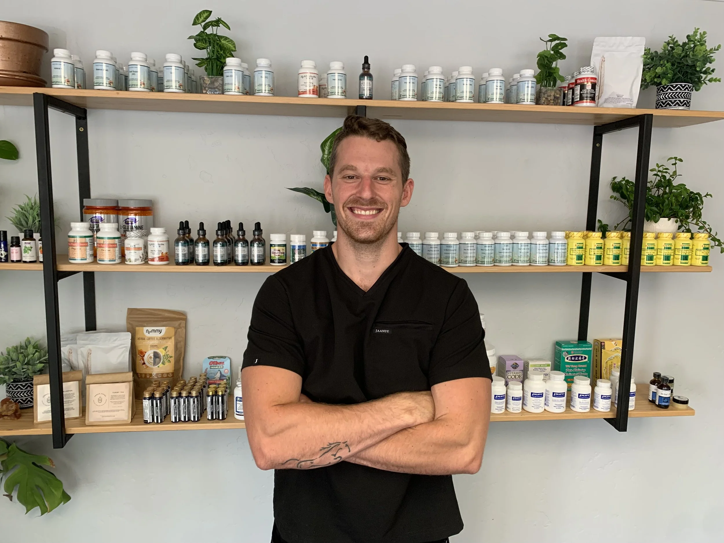 A smiling man in black standing with arms crossed in front of shelves filled with bottles and containers of supplements, vitamins, and herbal products in a health store or clinic.