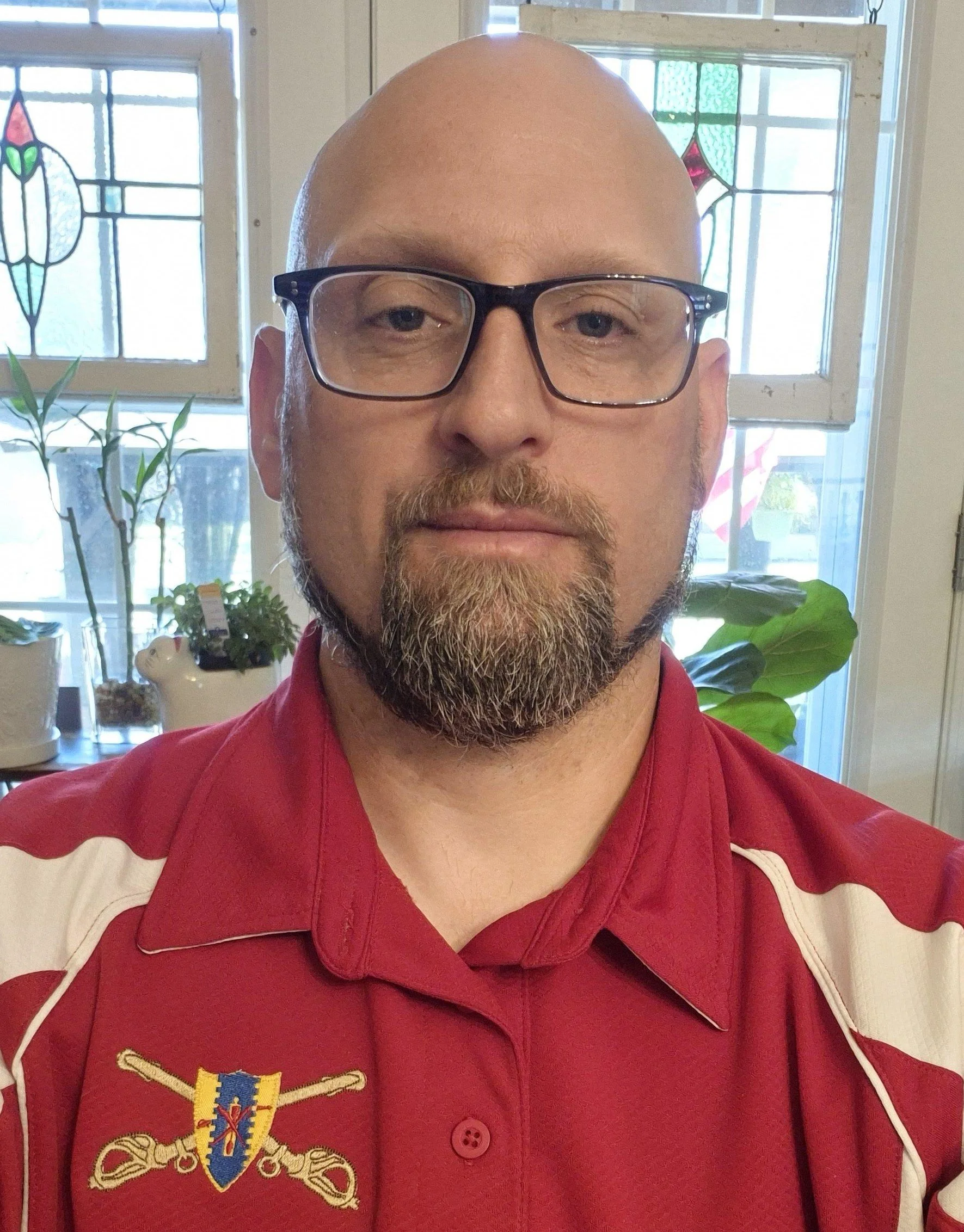 A man with glasses and a beard takes a selfie indoors, with stained glass windows and potted plants in the background.