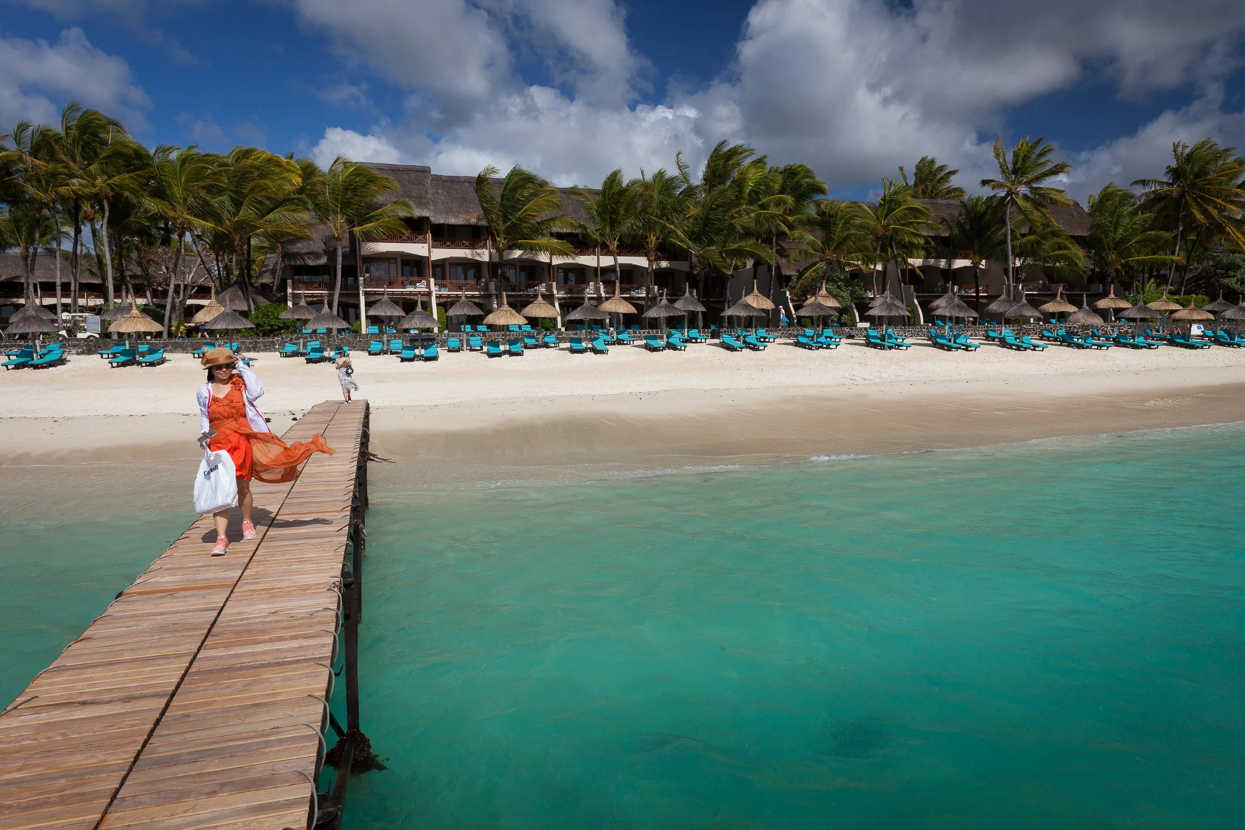 Woman in a flowing orange dress walking along the wooden jetty at Constance Belle Mare Plage, with the turquoise lagoon and beach loungers behind her.
