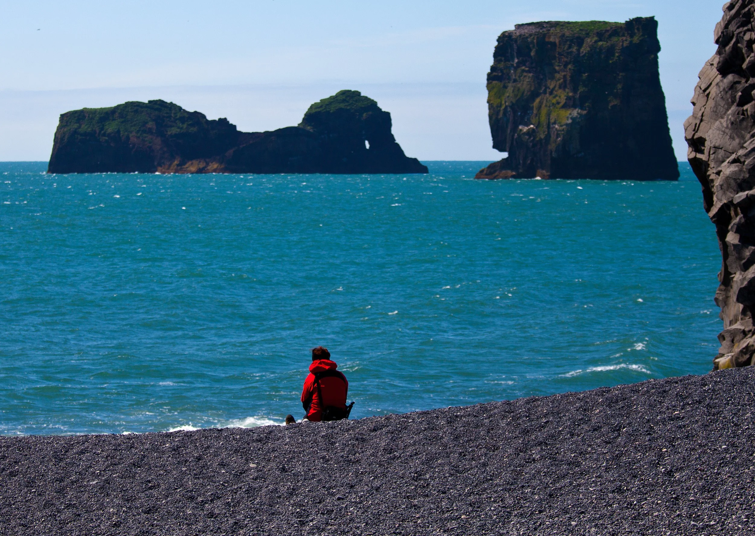 A lone figure on black sand, staring out at offshore sea stacks and the restless North Atlantic — Iceland’s south coast at its starkest.