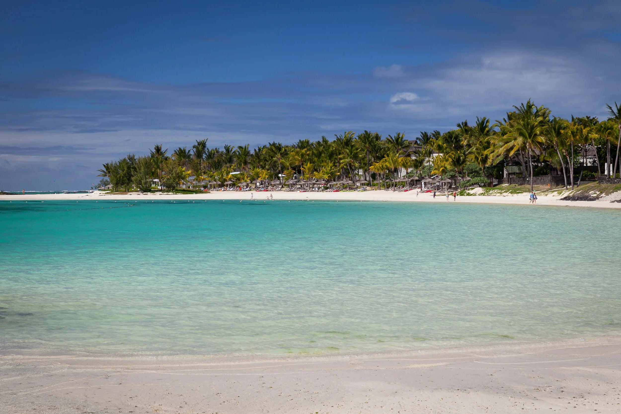 Long arc of Belle Mare Beach on Mauritius’ east coast, where shallow turquoise water meets powder-white sand and lines of palm trees.