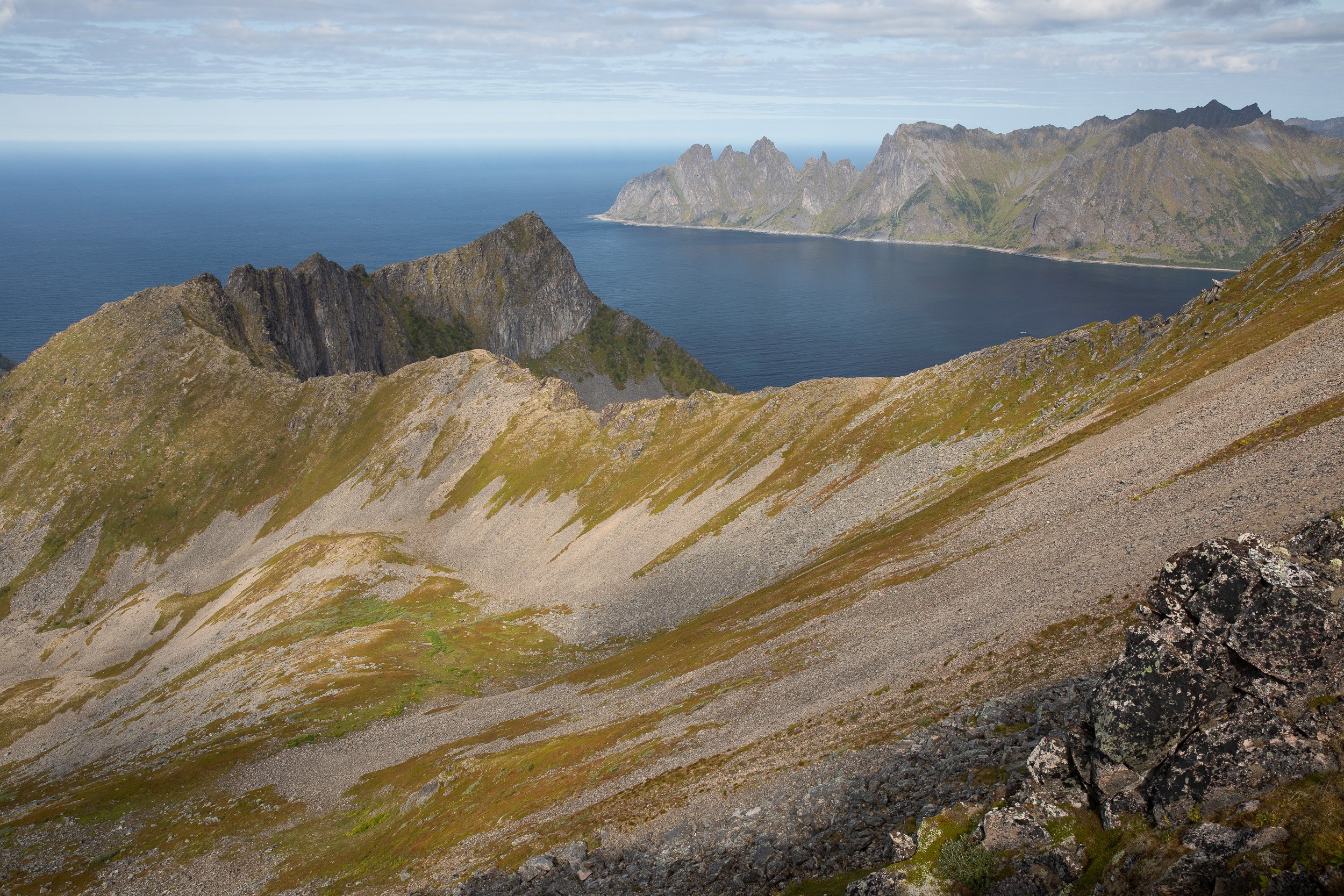 Rocky slopes below Husfjellet descend towards the open sea, with distant ridges and coastal mountains softened by shifting light.