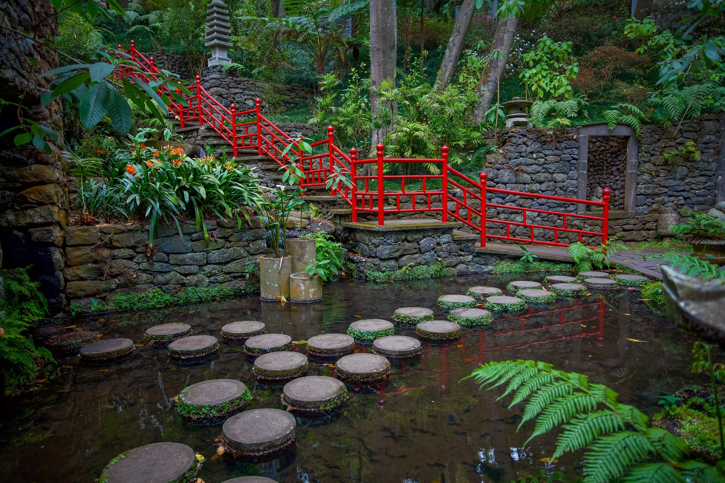 A red bridge and stepping stones lead through a lush Madeiran garden, reflected in still water and surrounded by ferns, stone walls and tropical planting.