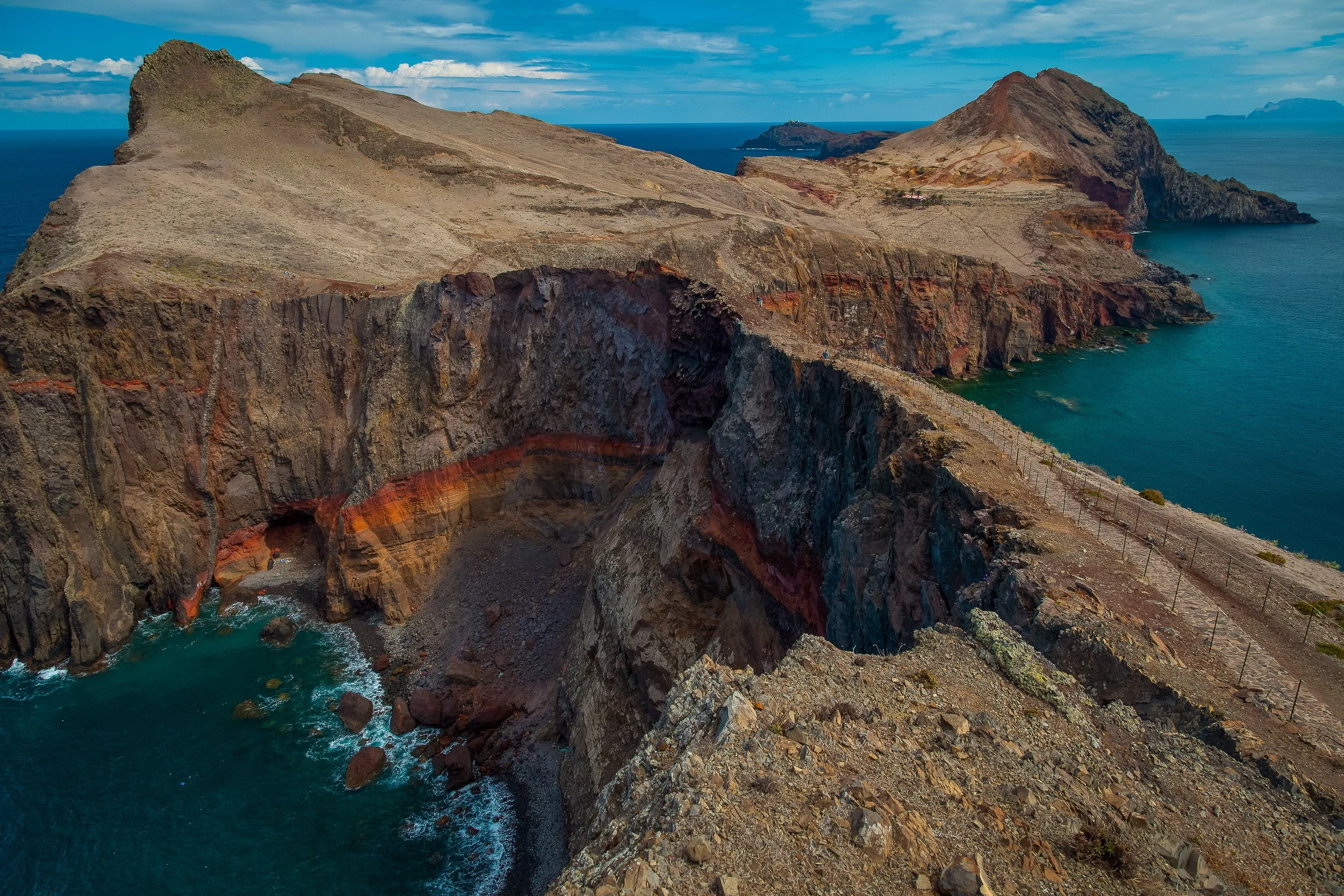 A wide view across Ponta de São Lourenço, where a narrow path follows the rugged volcanic ridge between cliffs, open sea and distant islands.