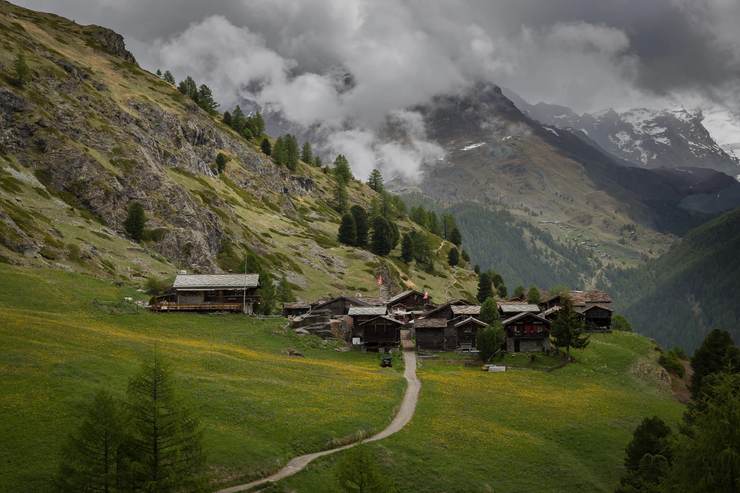 Cluster of old wooden barns and chalets on a steep alpine slope above Zermatt, overlooking deep valleys and distant mountains.