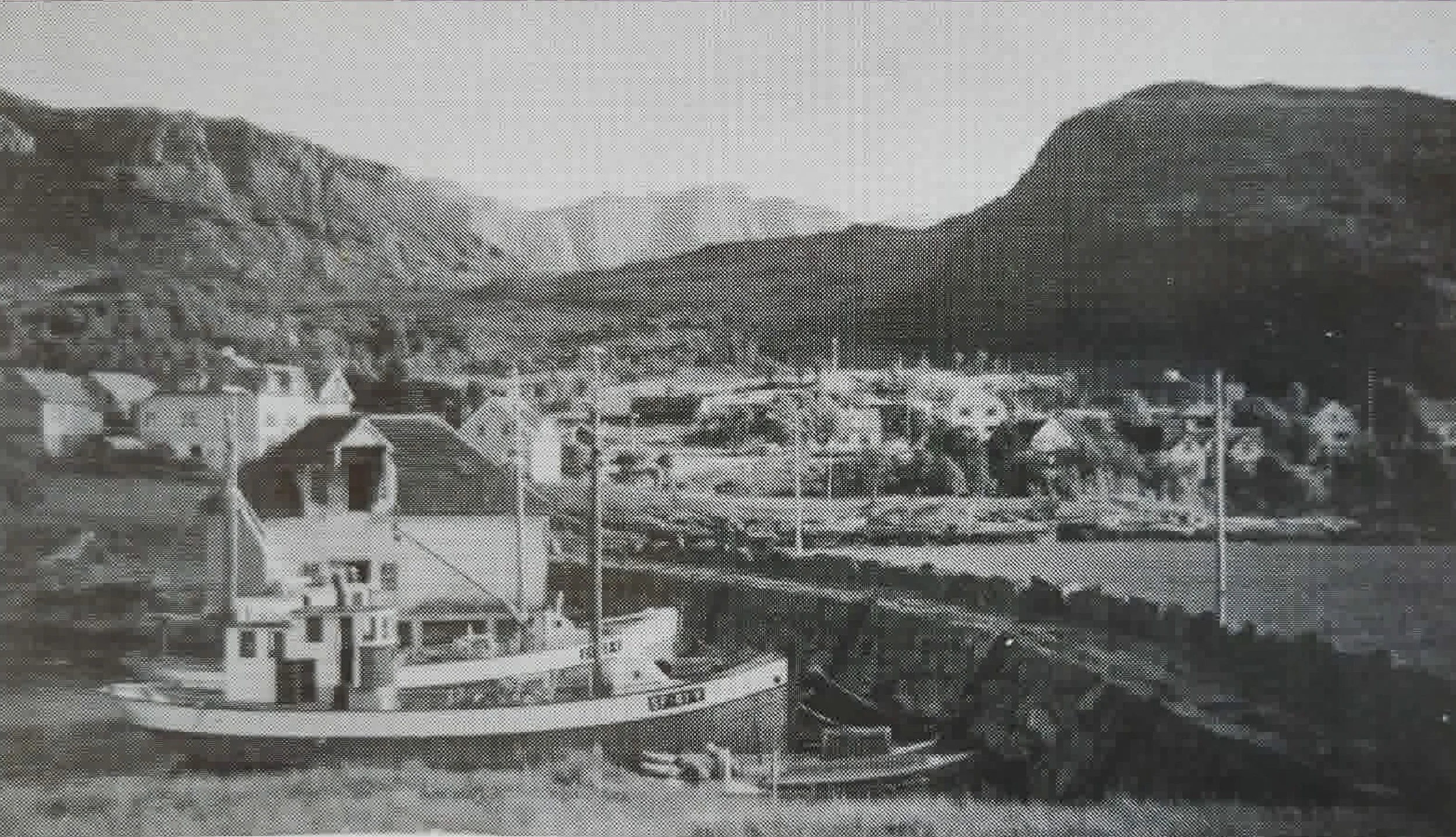 Svart-hvitt-bilde fra 1960 av motorkutteren «Strandholm» SF-61-D ved moloen i Husevåg, med hus og innmark i bakgrunnen.