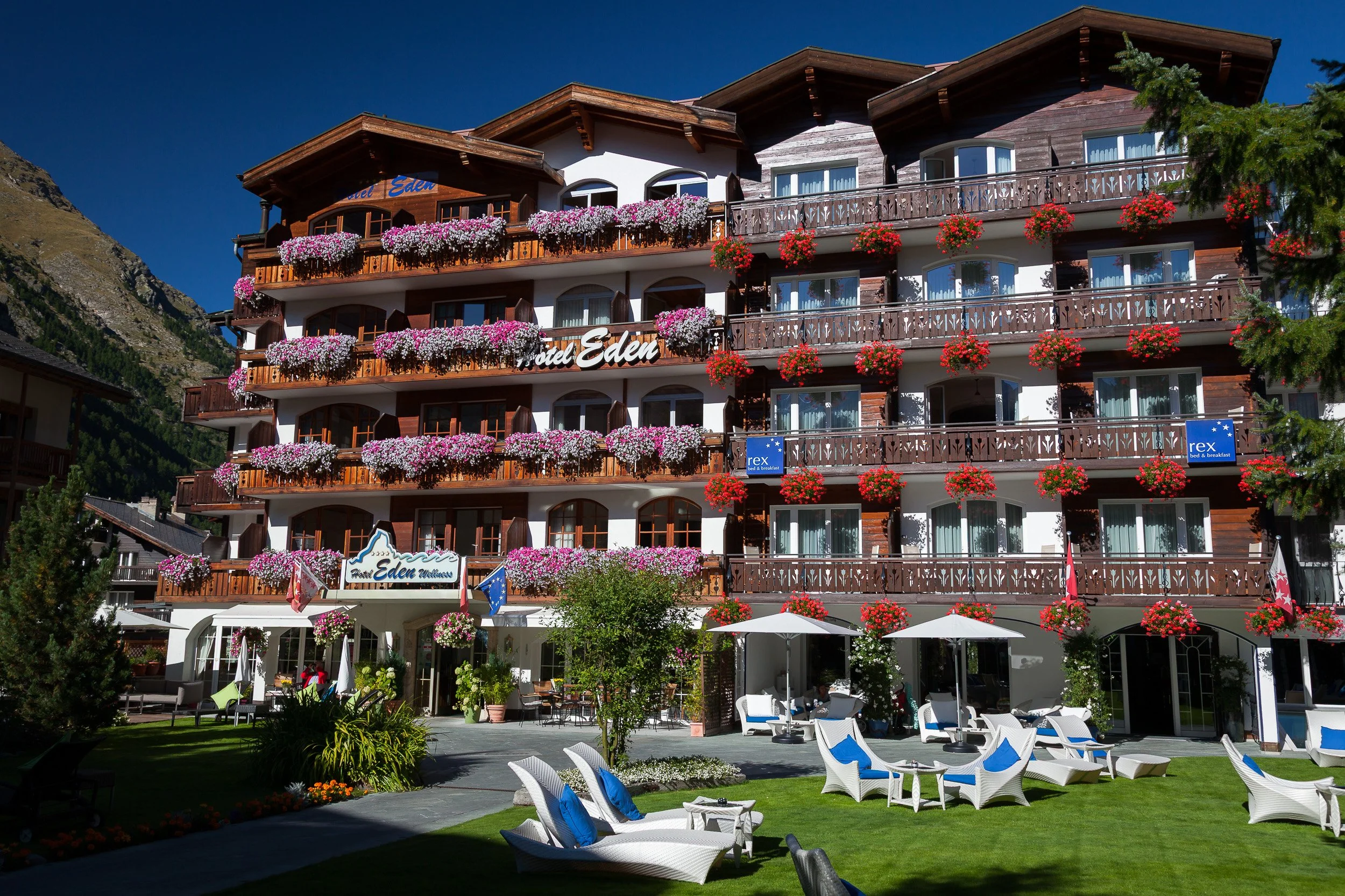 Hotel Eden’s sunlit garden in Zermatt with manicured lawn, loungers and balconies draped in cascades of pink and white flowers.