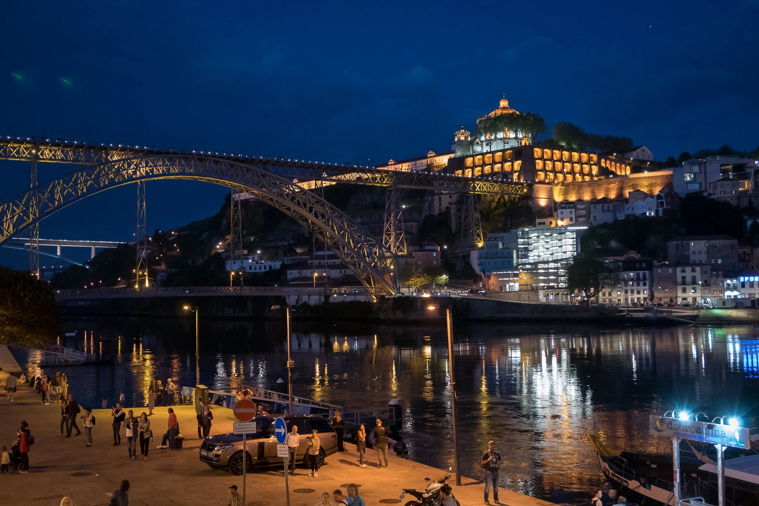 The Dom Luís I Bridge and Serra do Pilar are illuminated above the Douro River, reflected in the evening water from Vila Nova de Gaia.