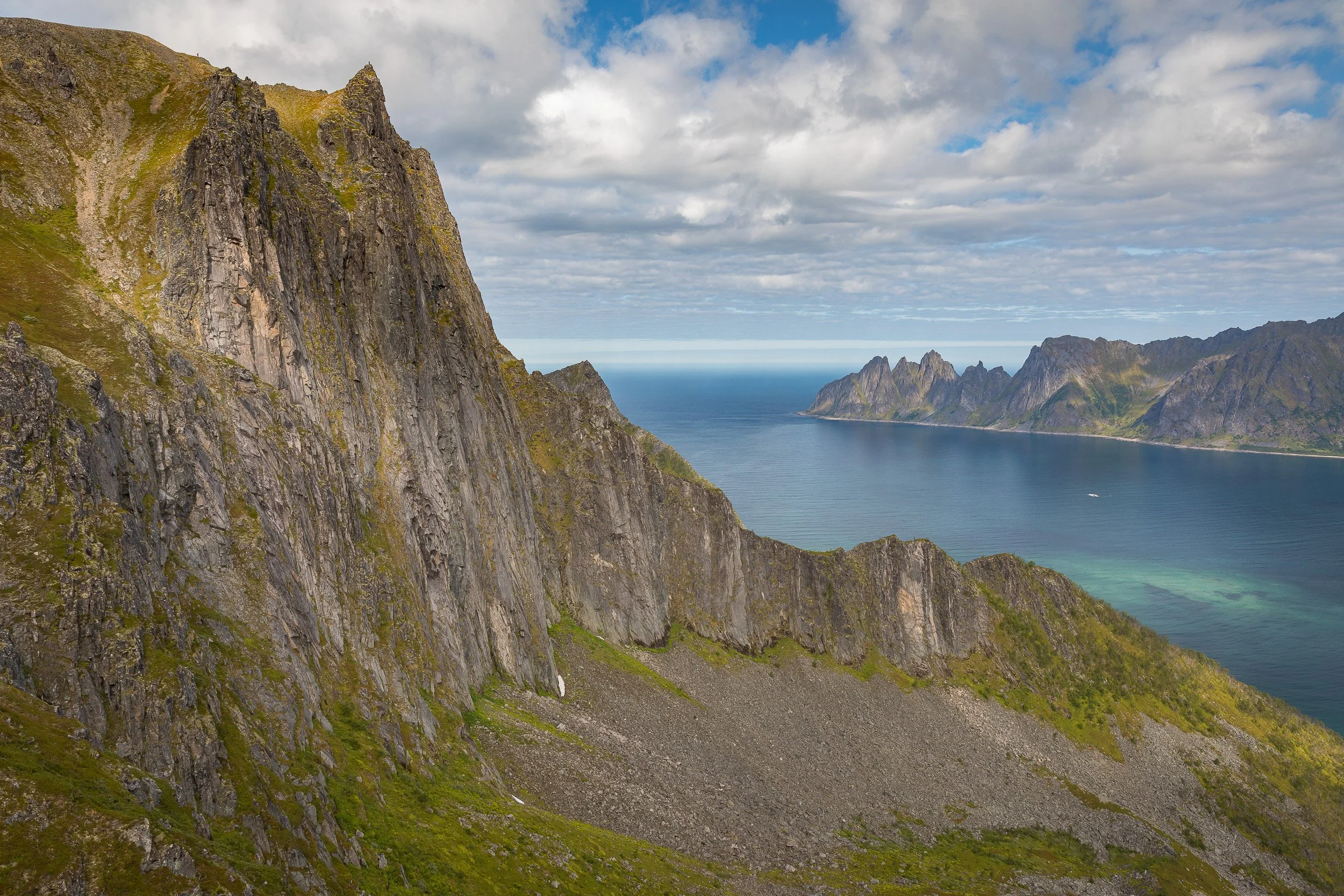 From Husfjellet, a steep rock wall falls towards the sea, with distant peaks and turquoise shallows opening across the Senja coast.