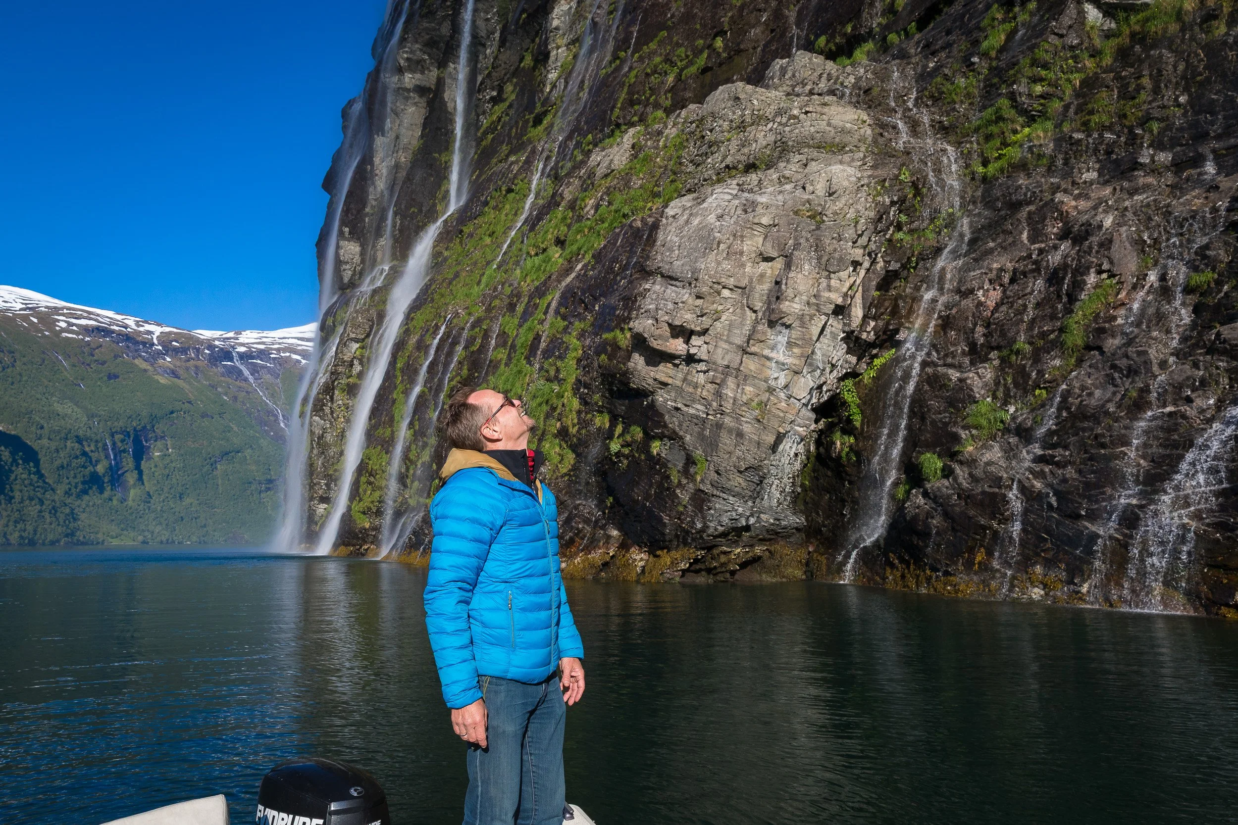 A man stands in the boat and looks up at the sheer, near-vertical mountain walls of Geirangerfjorden — scale that feels unreal.
