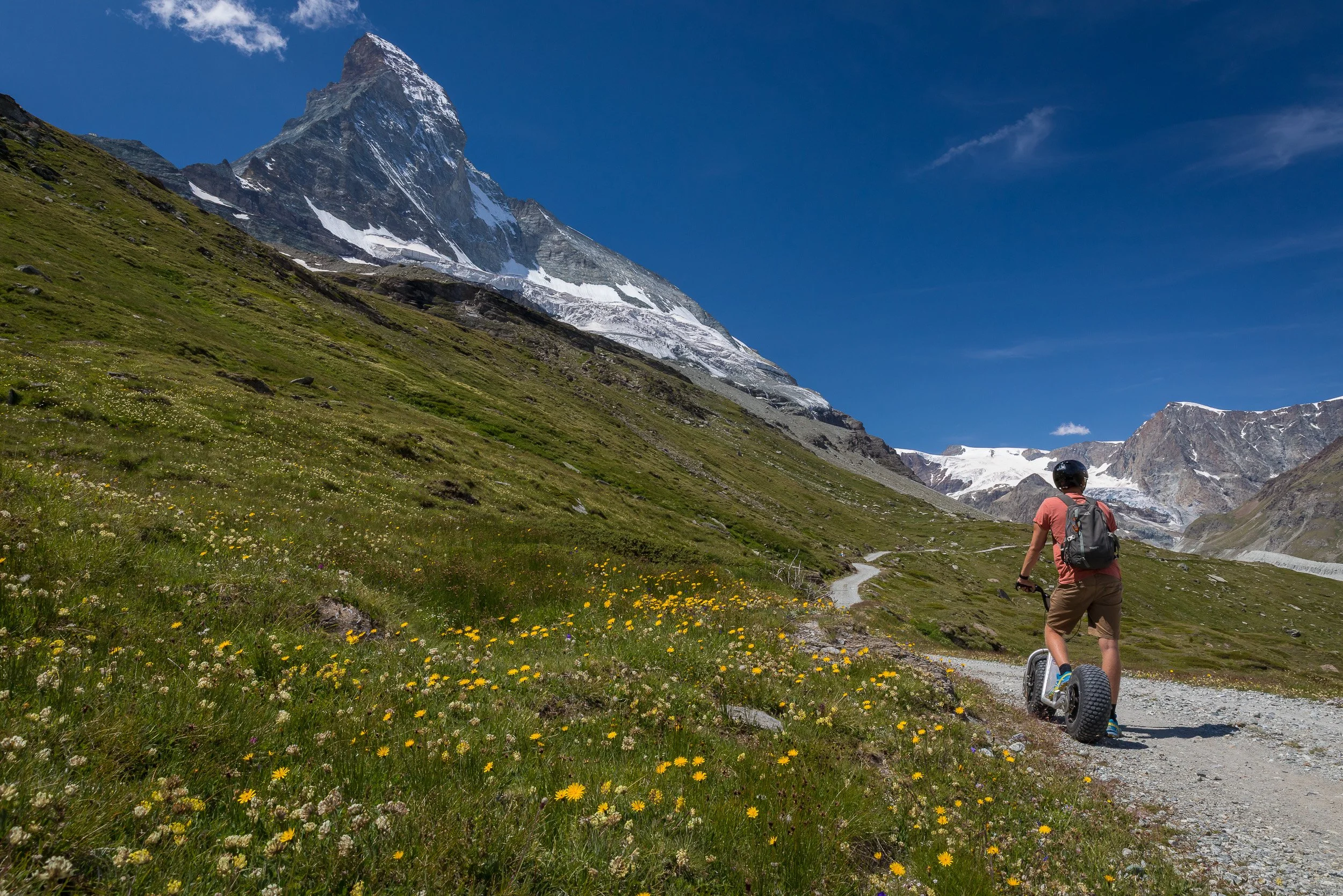 Young rider on an off-road scooter following a winding alpine trail towards the Matterhorn through a meadow of yellow wildflowers.