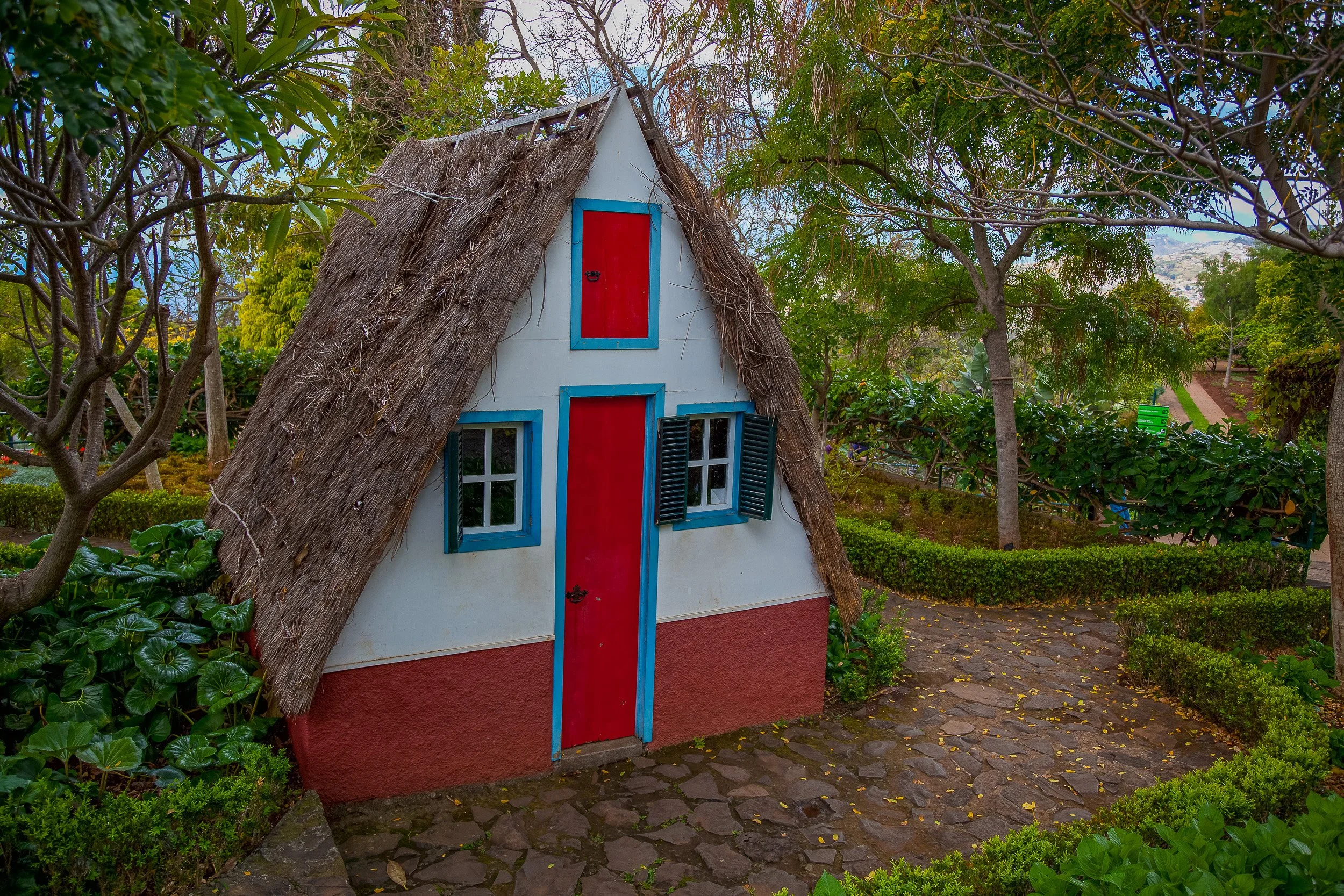 A traditional Madeiran thatched house set among garden paths and trees, with red doors and blue trim adding colour to the quiet green setting.