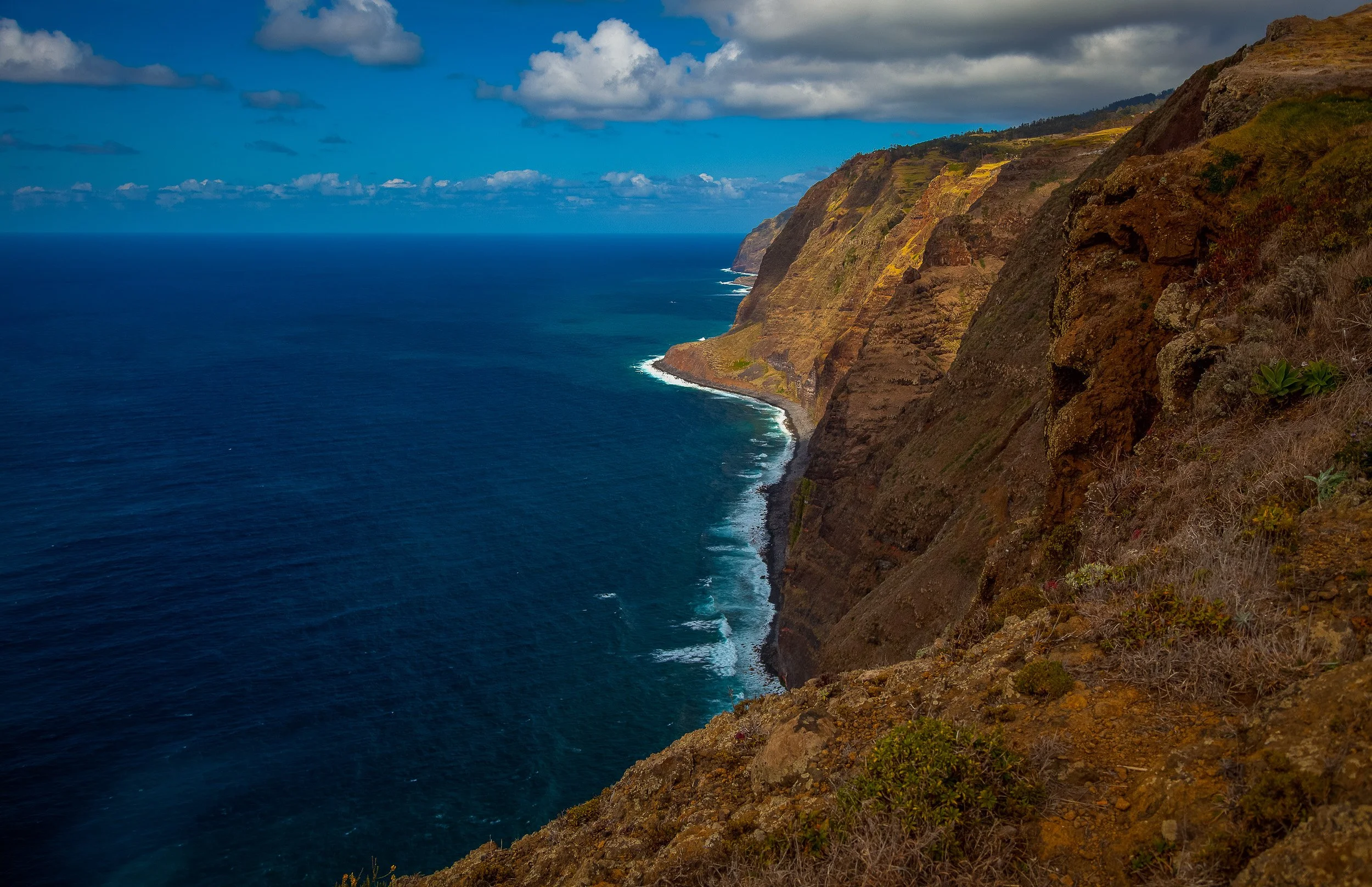 A dramatic sweep of Madeira’s west coast near Ponta do Pargo, with layered cliffs, turquoise surf and low clouds casting shade across the Atlantic edge.