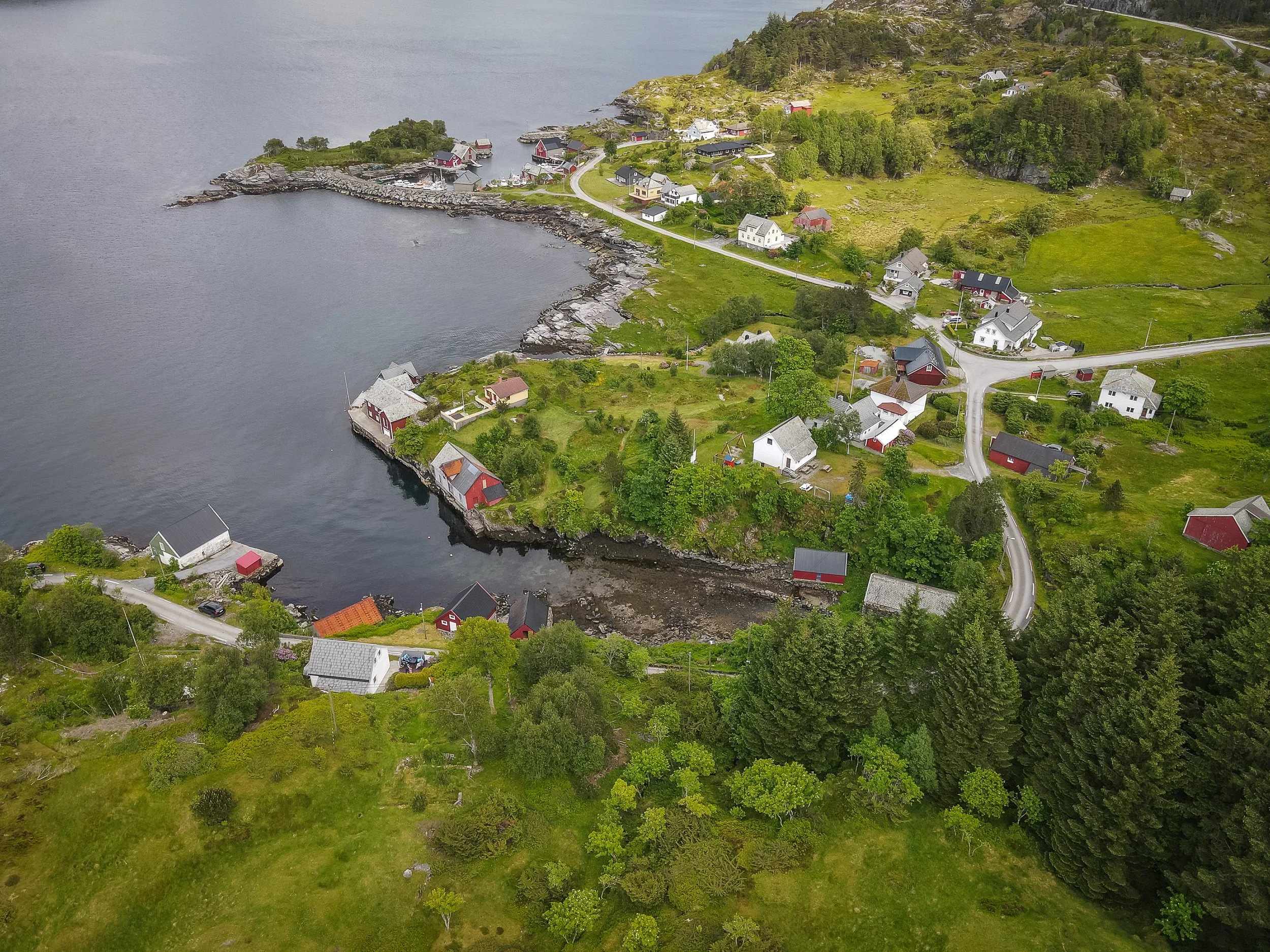 Luftfoto av Husevåg på Husevågøy, med en lun våg, sjøhus, naust, spredt bebyggelse, veier, grønn innmark og fjorden rundt bygda.