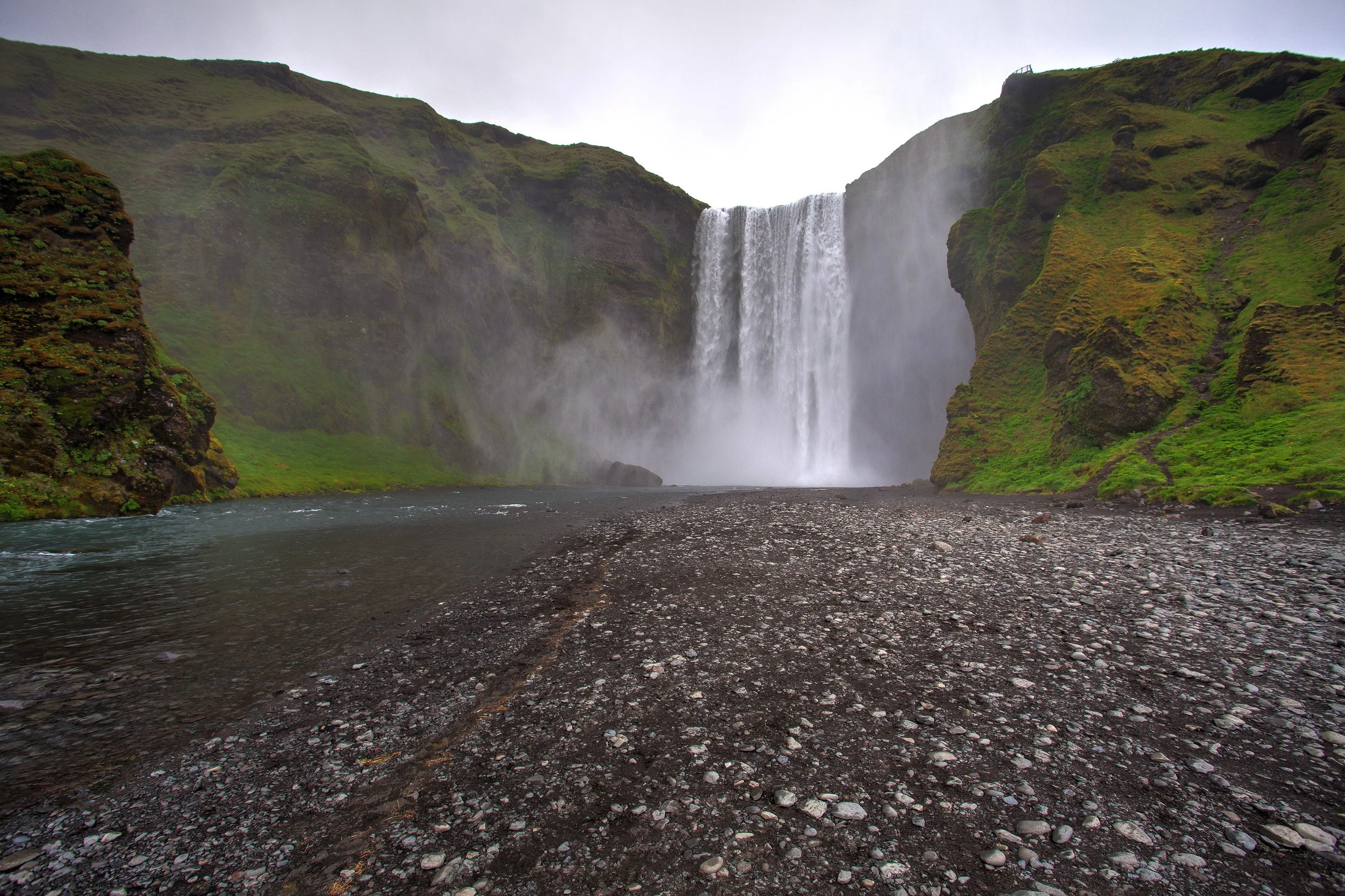 Skógafoss — the waterfall thunders into mist at the end of a stony riverbed, framed by mossy cliffs and a pale, rain-washed sky.