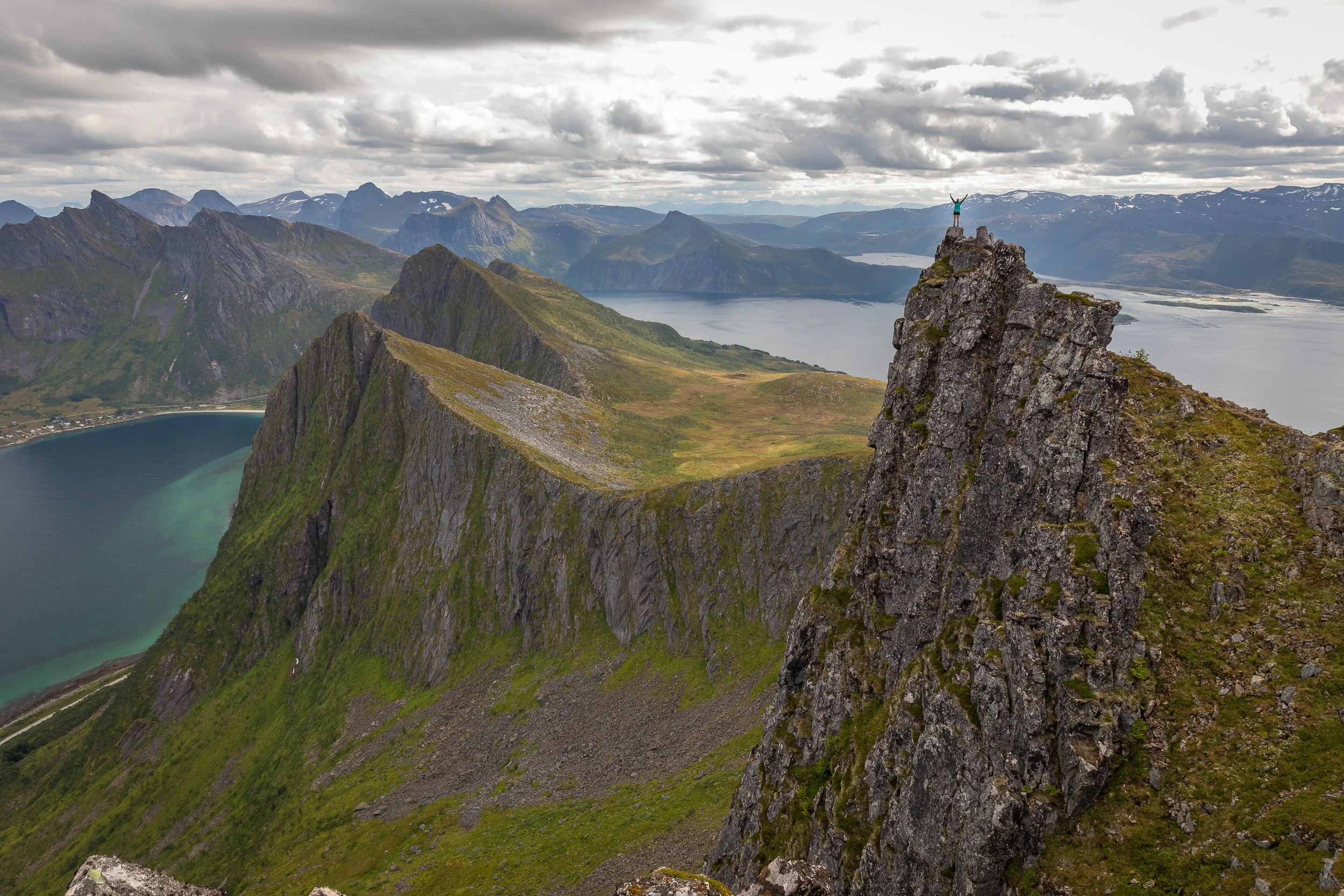 A lone hiker stands on a rocky outcrop below Husfjellet, surrounded by sweeping ridges, fjords and cloud-shadowed mountains.