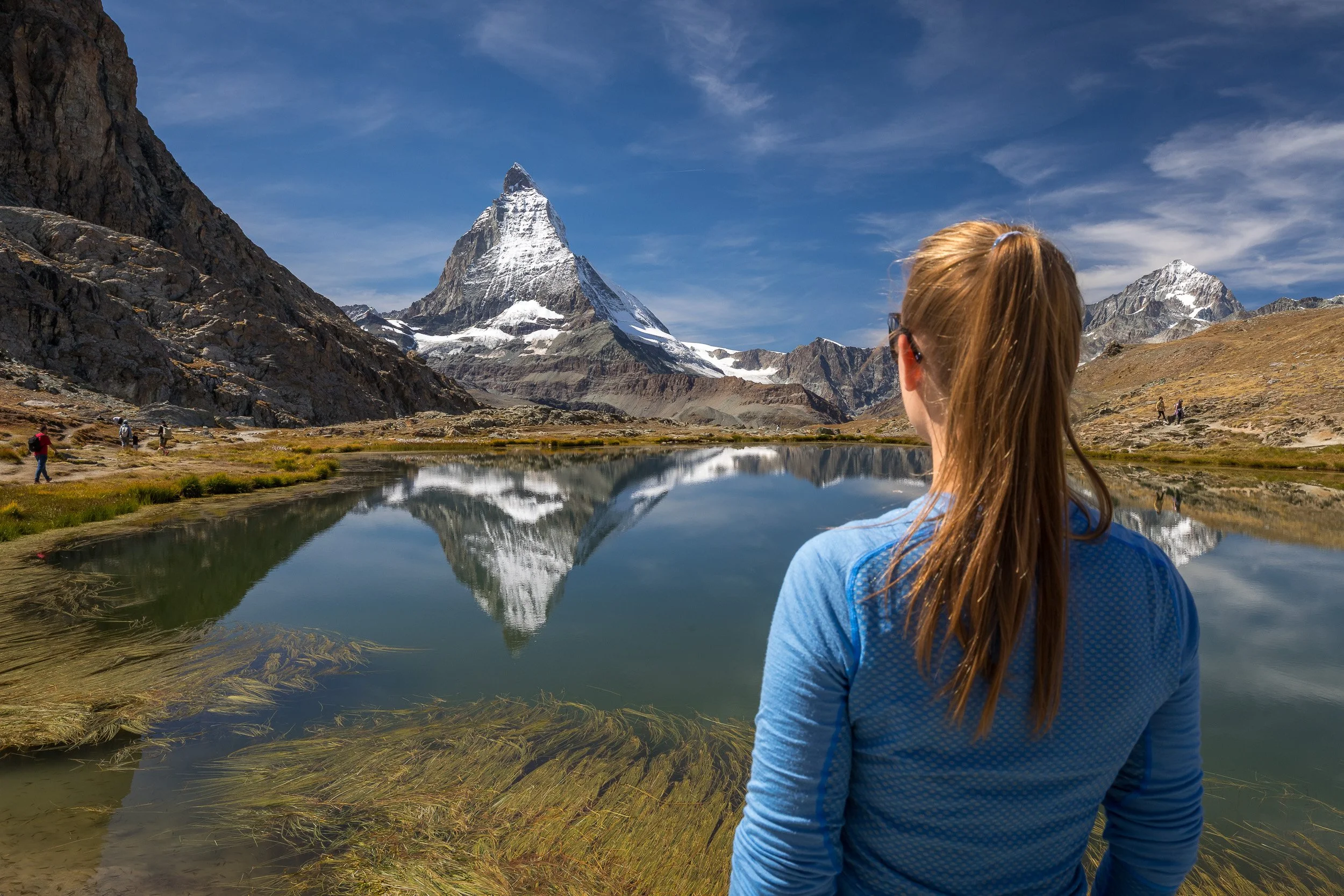 Woman standing beside Riffelsee, watching the mirror-like reflection of the Matterhorn and the surrounding alpine peaks.
