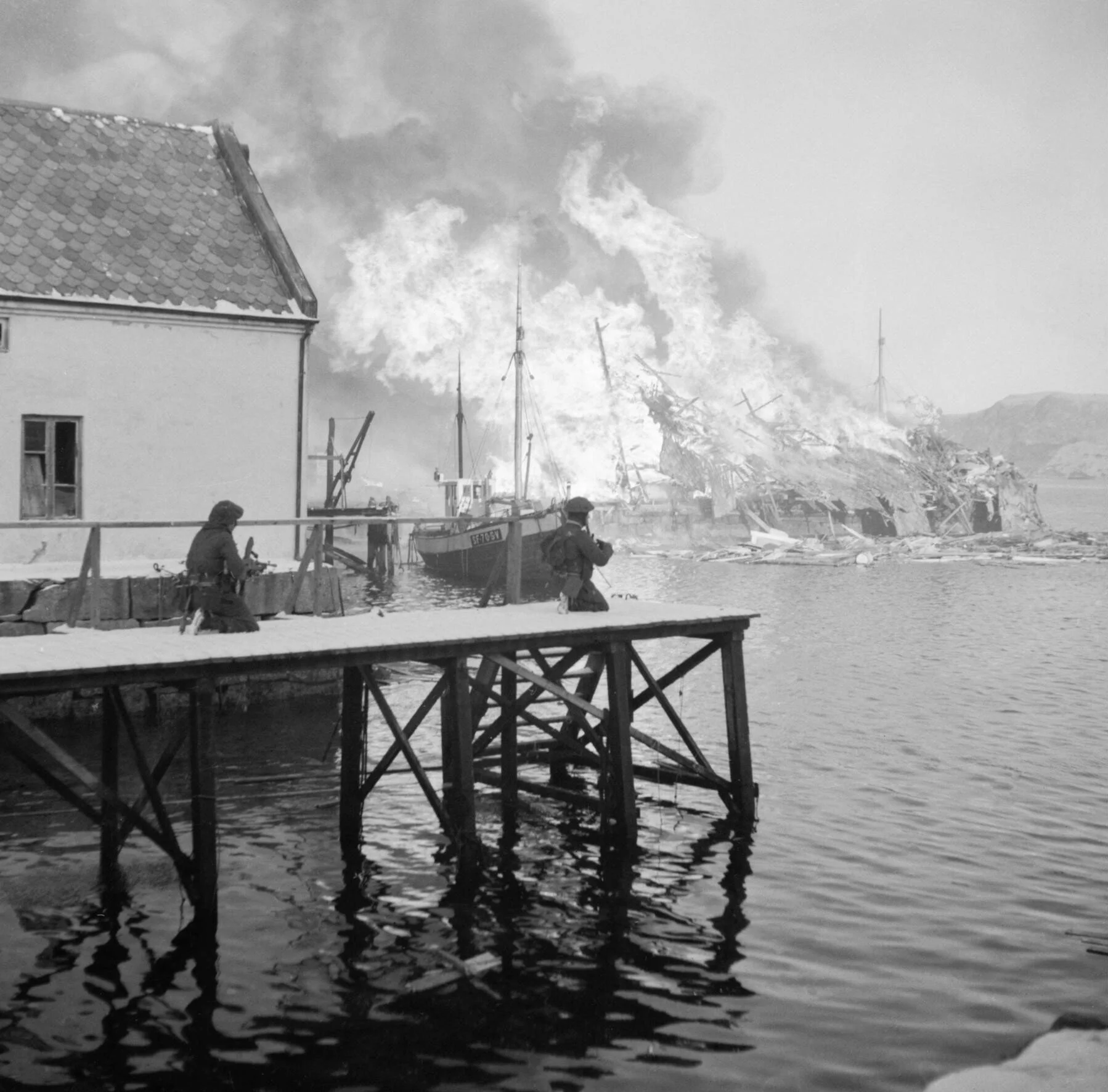 Historisk bilde fra Måløyraidet 27. desember 1941 med brann ved en oljefabrikk og britiske soldater på kaien.