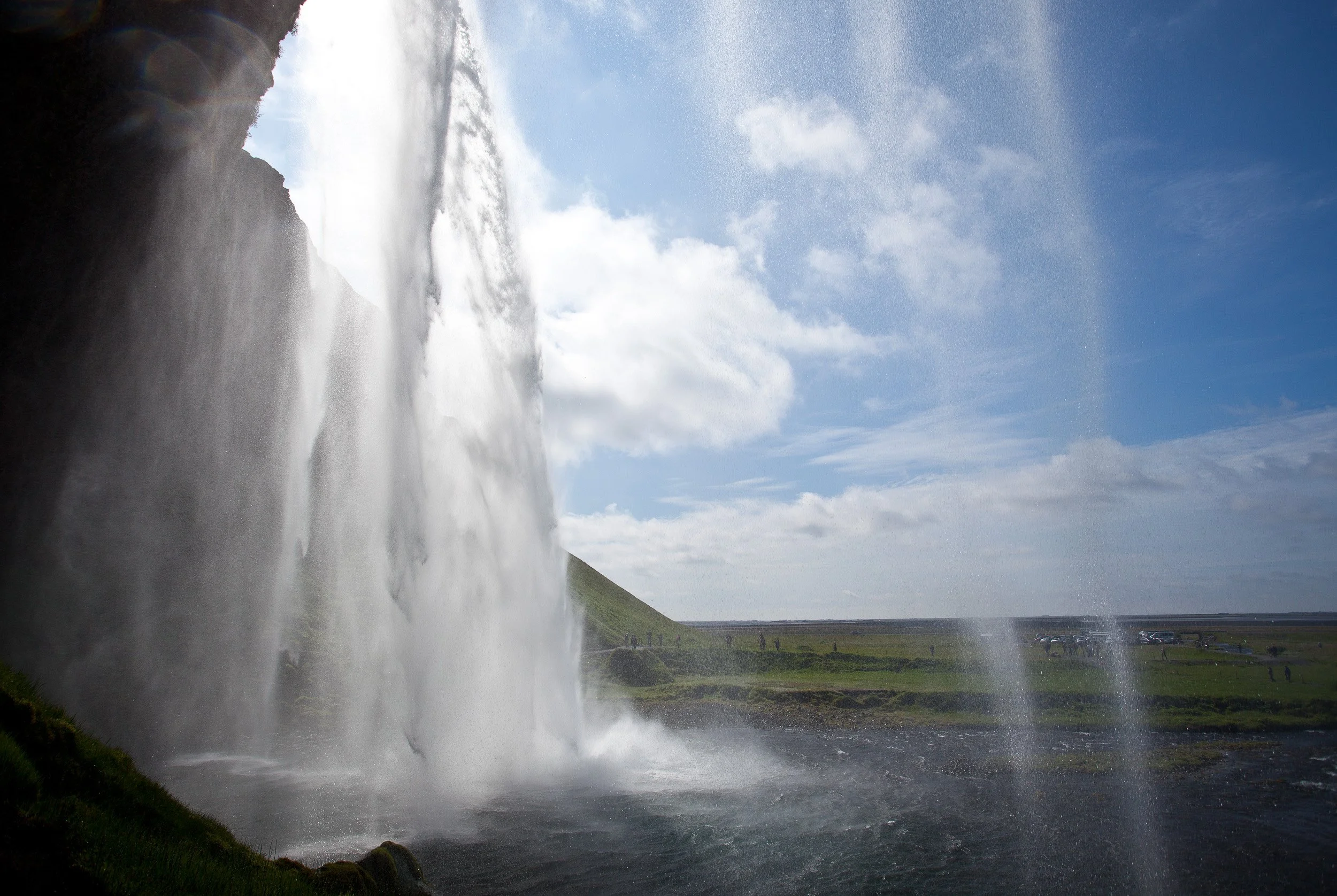 Behind Seljalandsfoss — standing in the shadowed cave as the waterfall falls like a curtain, the landscape beyond blurred by drifting spray.