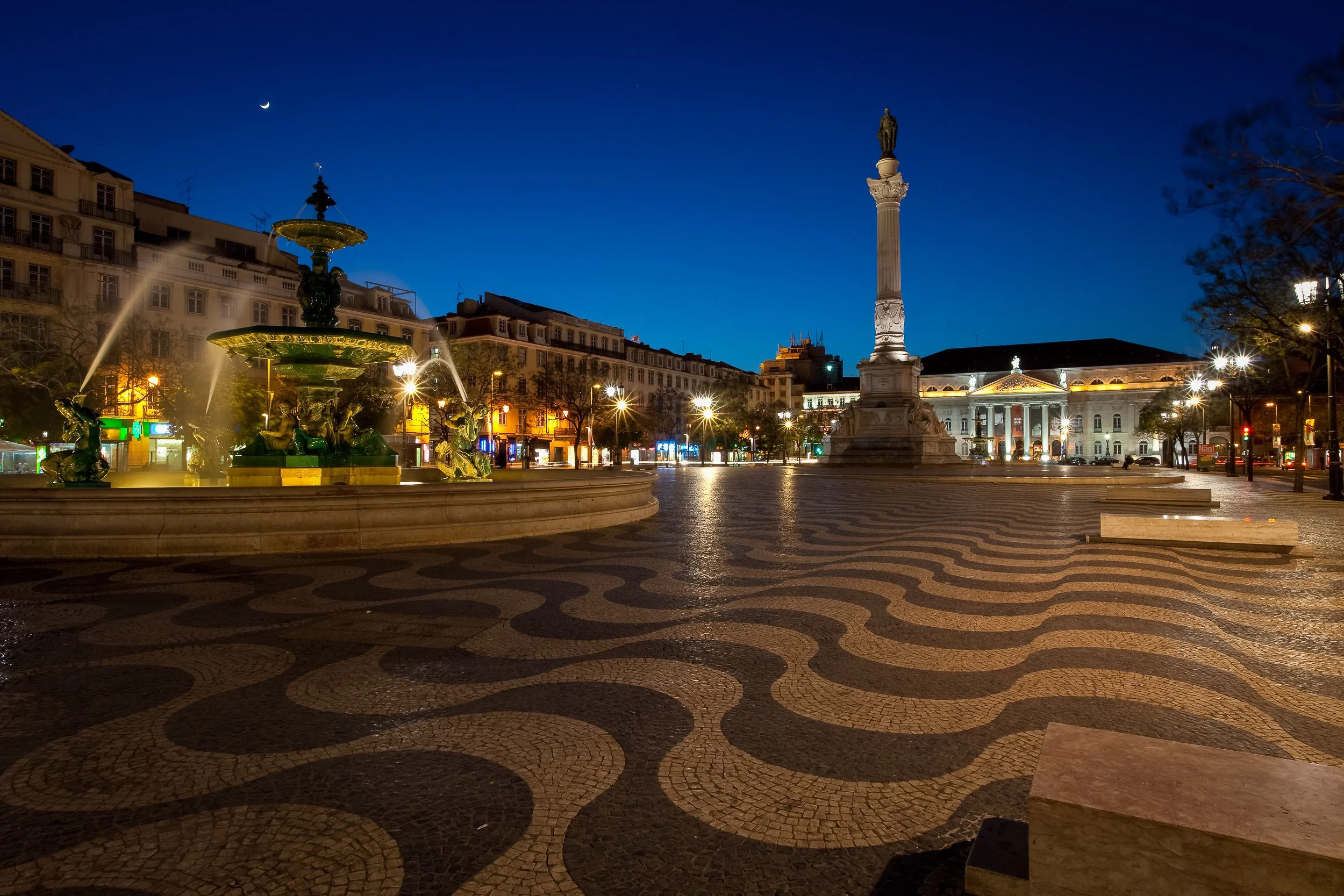 Rossio Square in Lisbon after dark, with the fountain, wave-patterned pavement and Pedro IV Column lit against the evening sky.