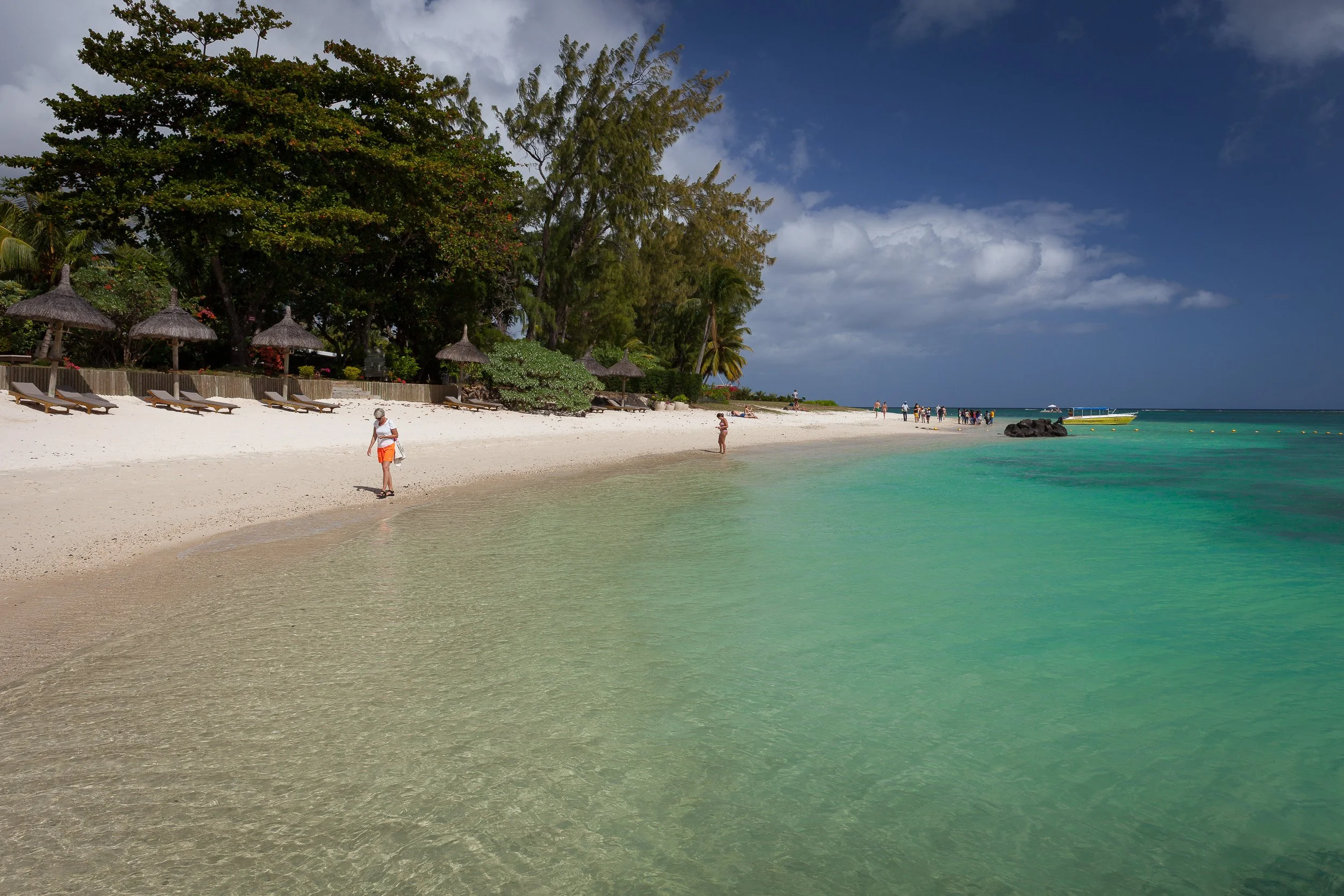 People strolling along the soft sand at Trou aux Biches beach in Grand Baie, with thatched parasols and clear emerald water.