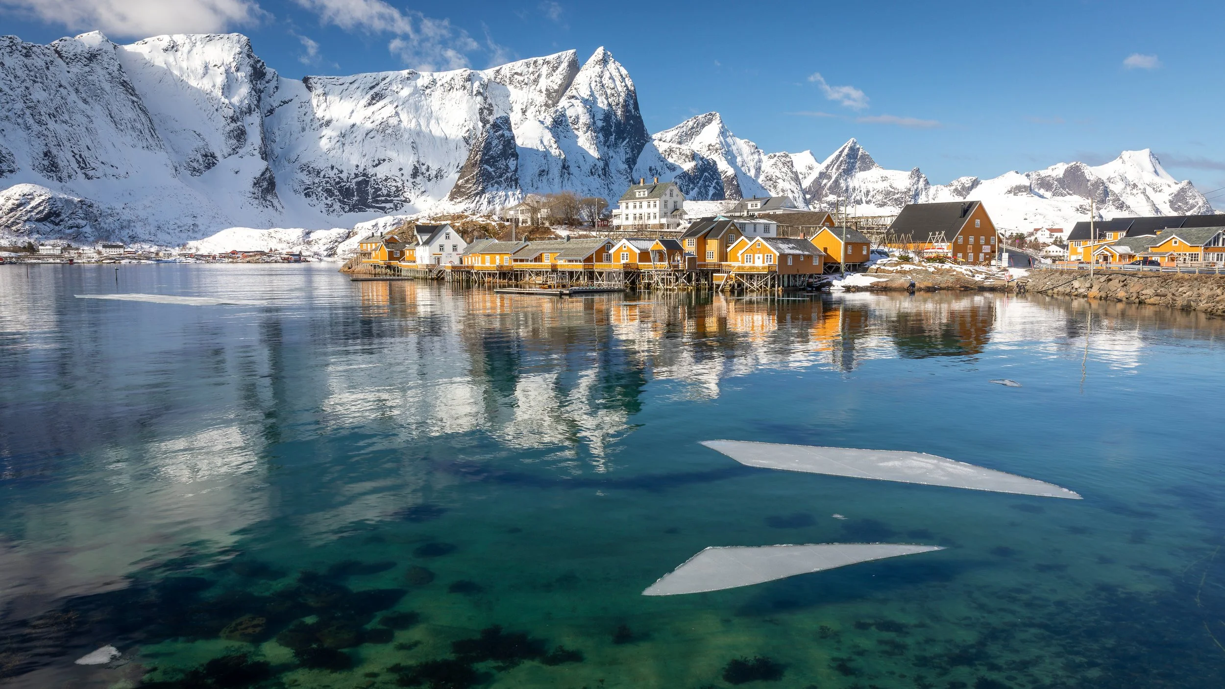 Orange rorbuer by calm winter water in Hamnøy, with snow-covered peaks rising behind in Lofoten, Norway.