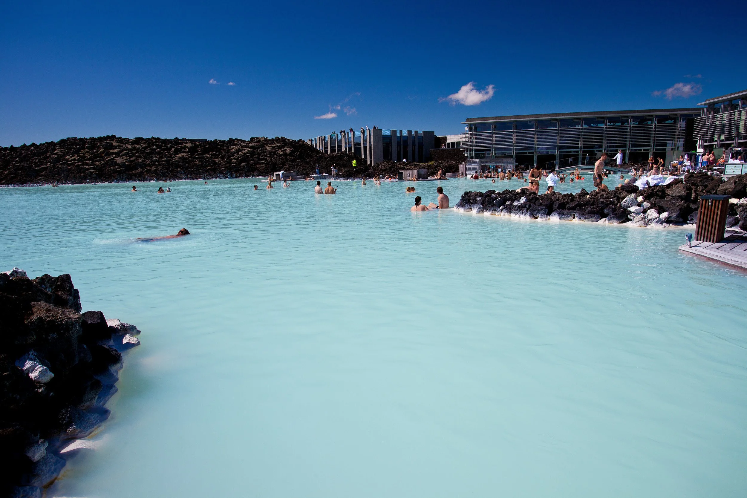 The milky-turquoise water of the Blue Lagoon stretches between jagged black lava rocks, with bathers drifting in the steam under a deep Icelandic blue sky
