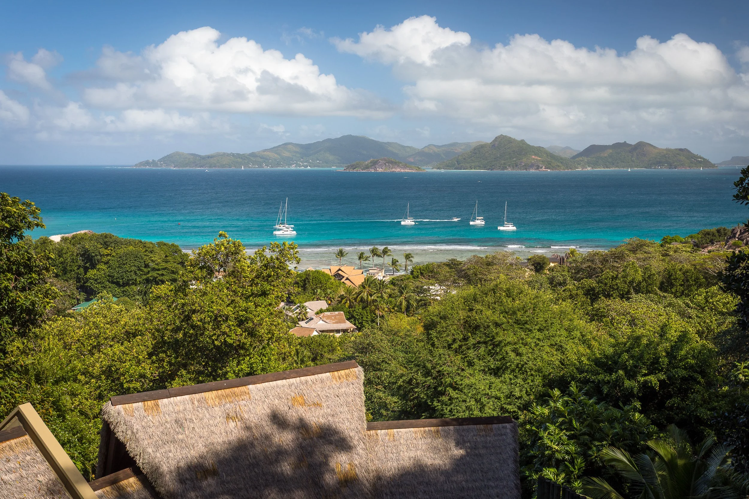 View from La Digue towards Praslin – turquoise lagoon and sailing catamarans beyond palm-fringed rooftops, with green hills under a bright sky.