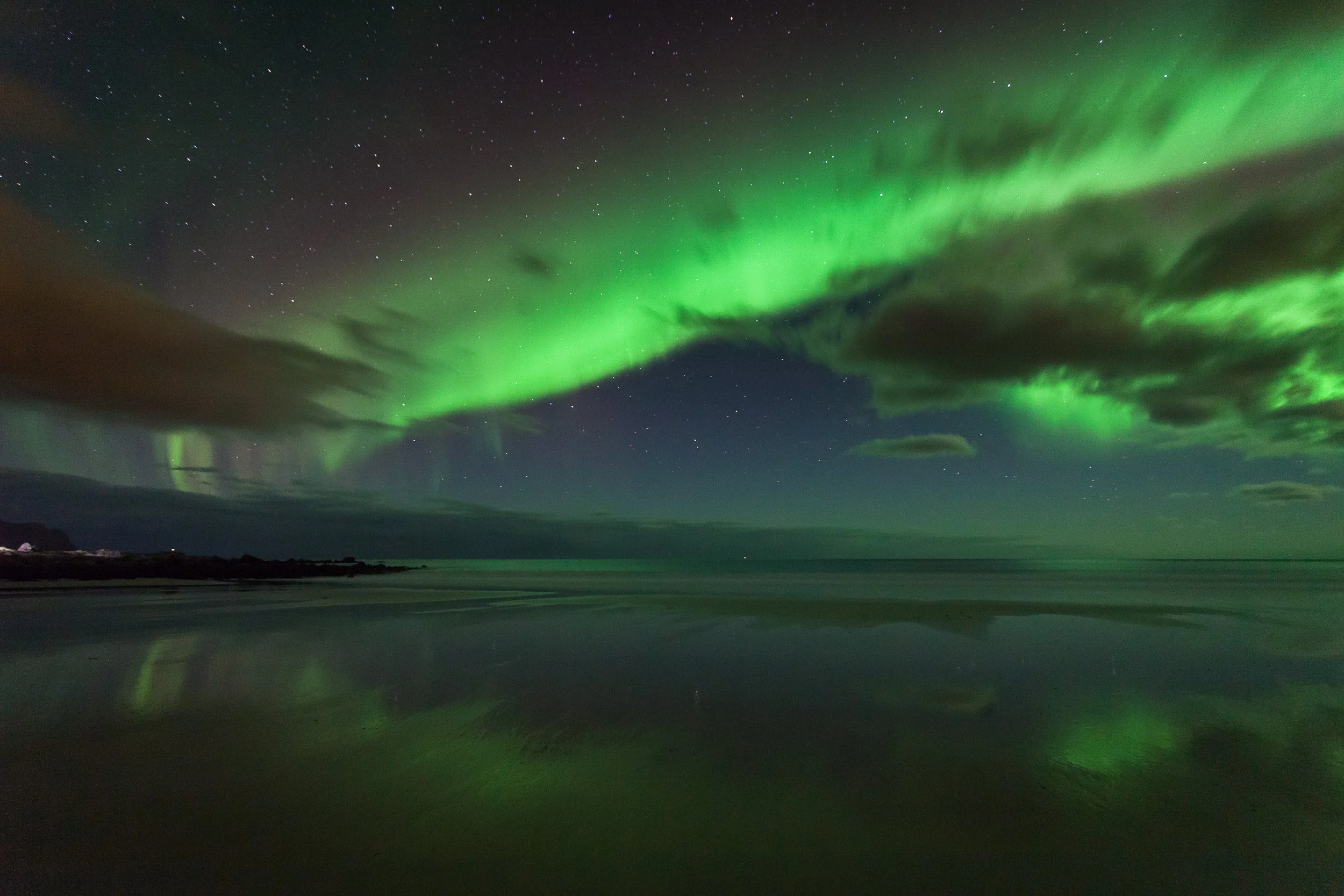 Aurora borealis over Skagsanden beach at Flakstad in Lofoten, with the northern lights reflected in the wet sand and shallow water along the Arctic shoreline.