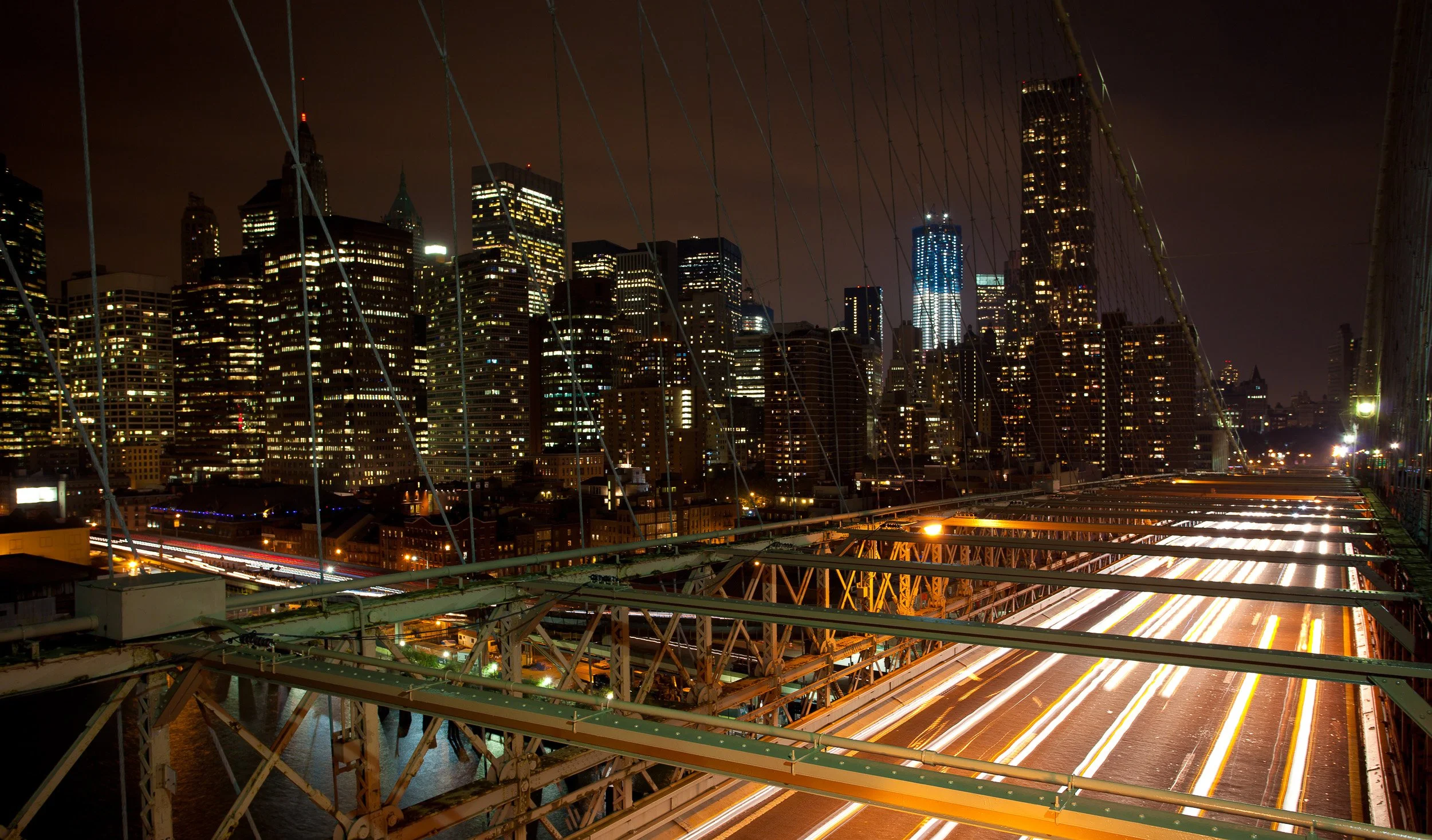 Night traffic streaming across the Brooklyn Bridge with Lower Manhattan blazing behind.