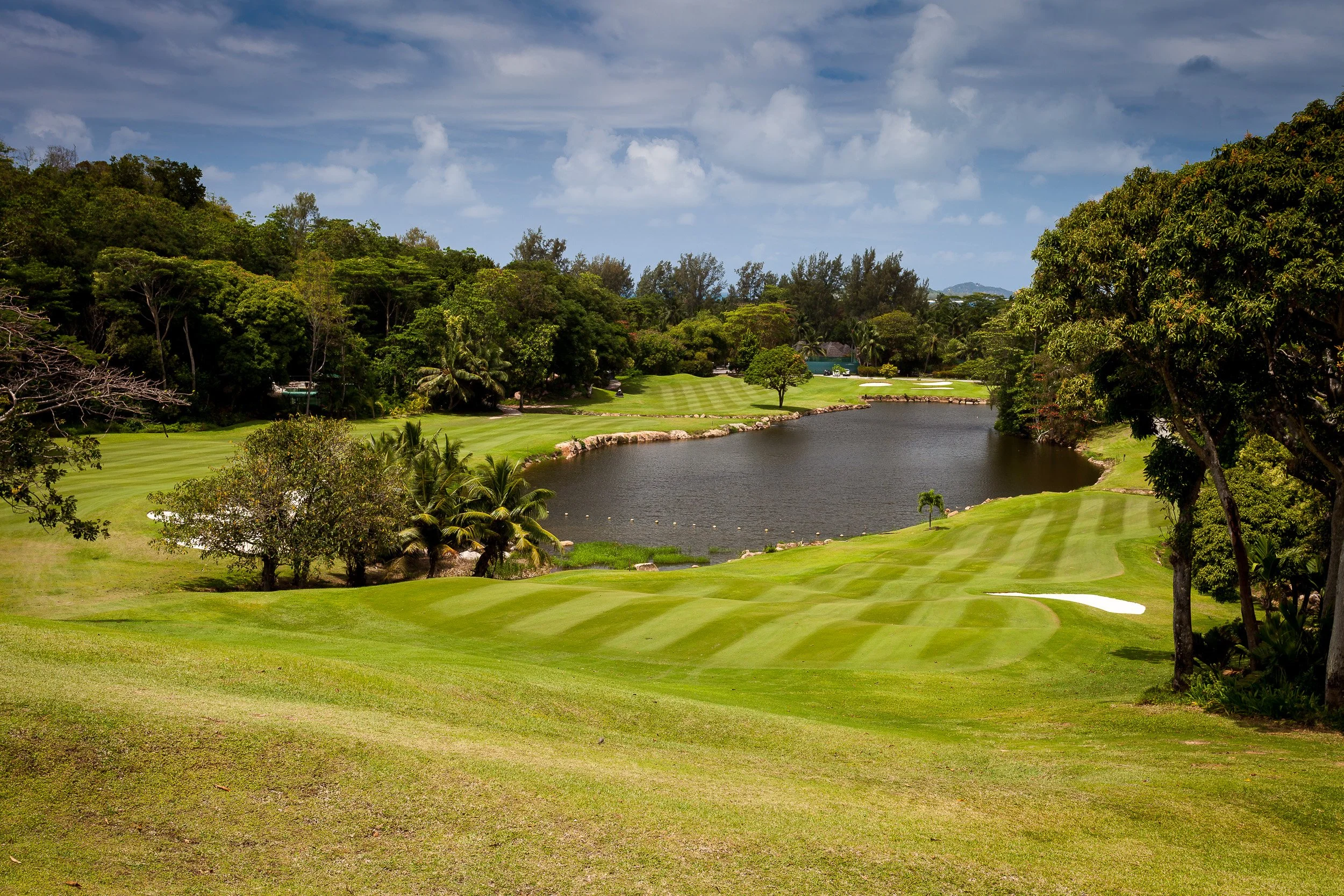 Constance Lémuria golf course on Praslin – lush fairways curving around a small lagoon, framed by palm trees, forest and a partly cloudy tropical sky.