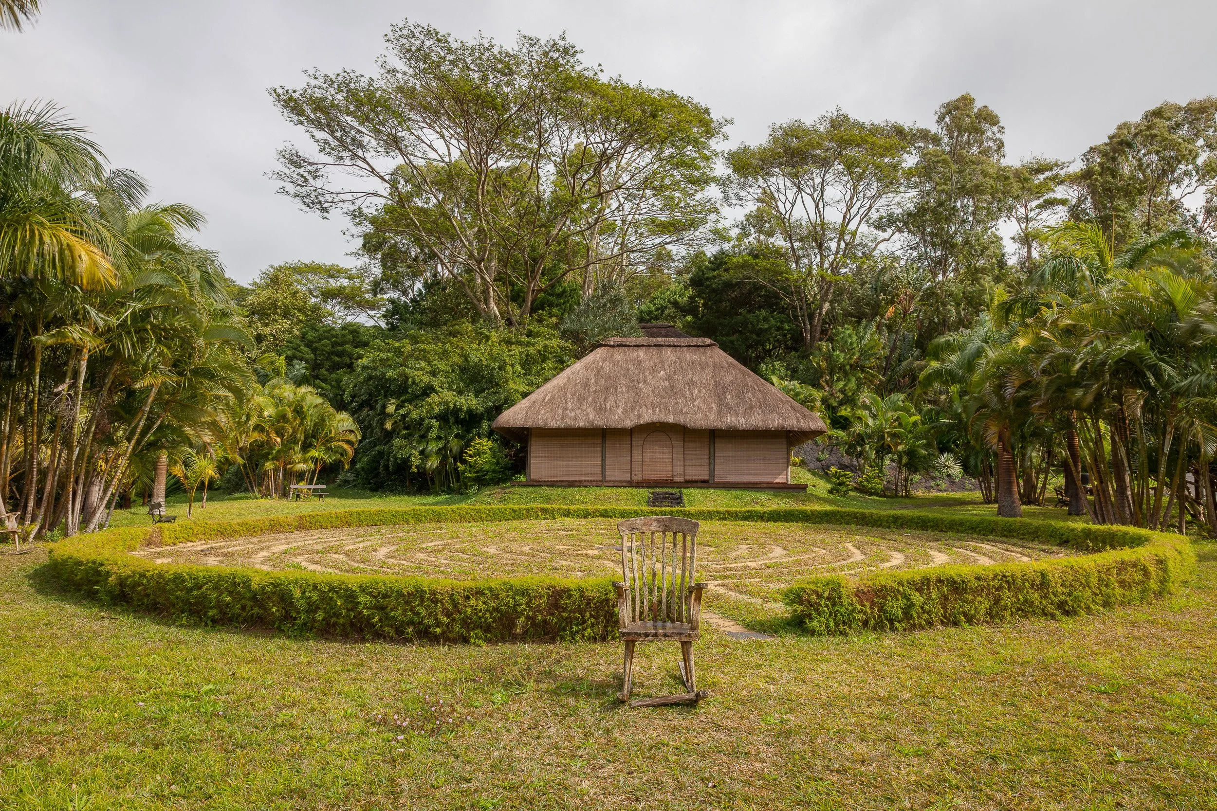 Wooden chair facing a circular garden labyrinth and thatched meditation pavilion at Lakaz Chamarel, surrounded by lush green hills.