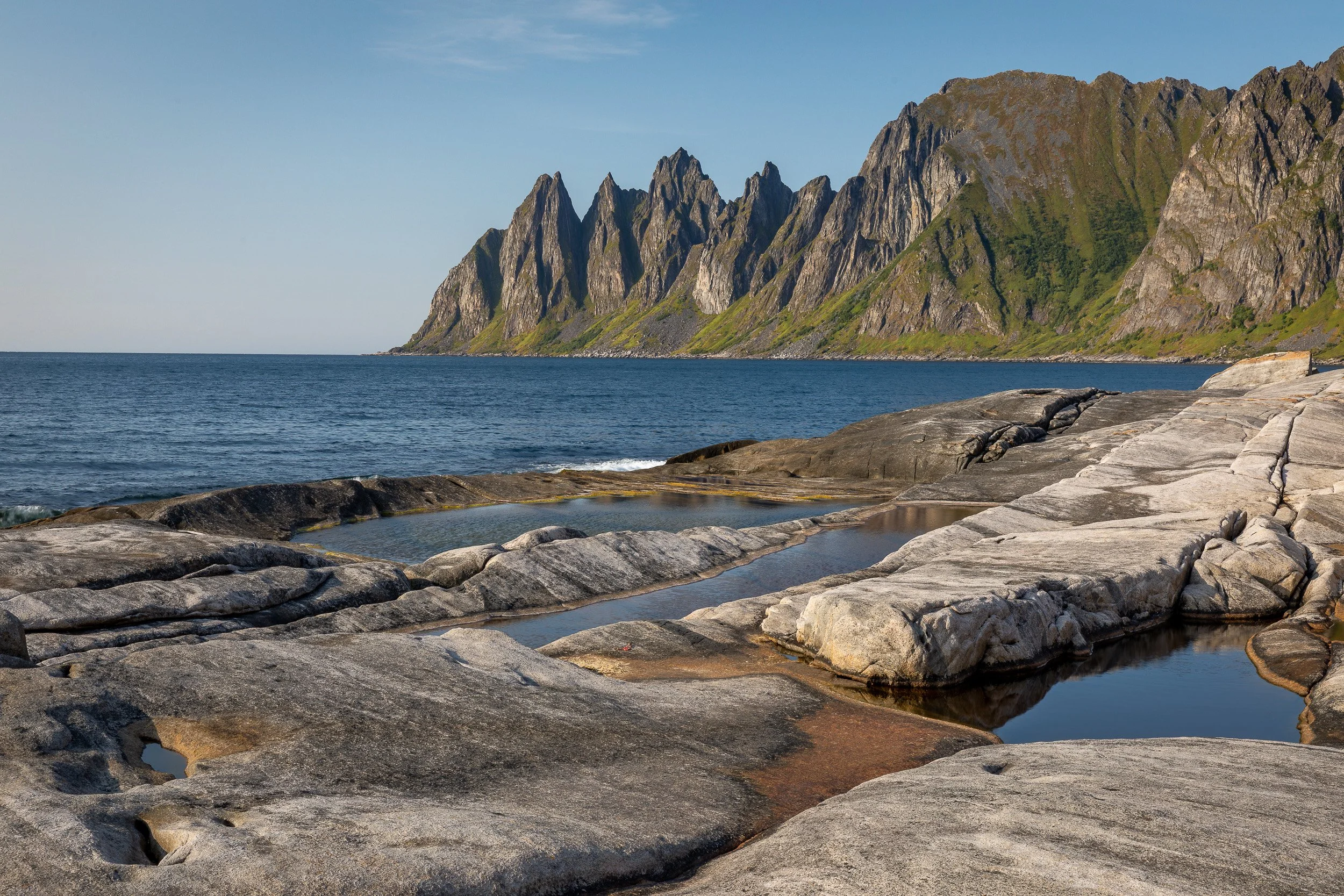 Smooth coastal rocks and still pools at Tungeneset, looking towards the jagged Okshornan peaks across the sea.