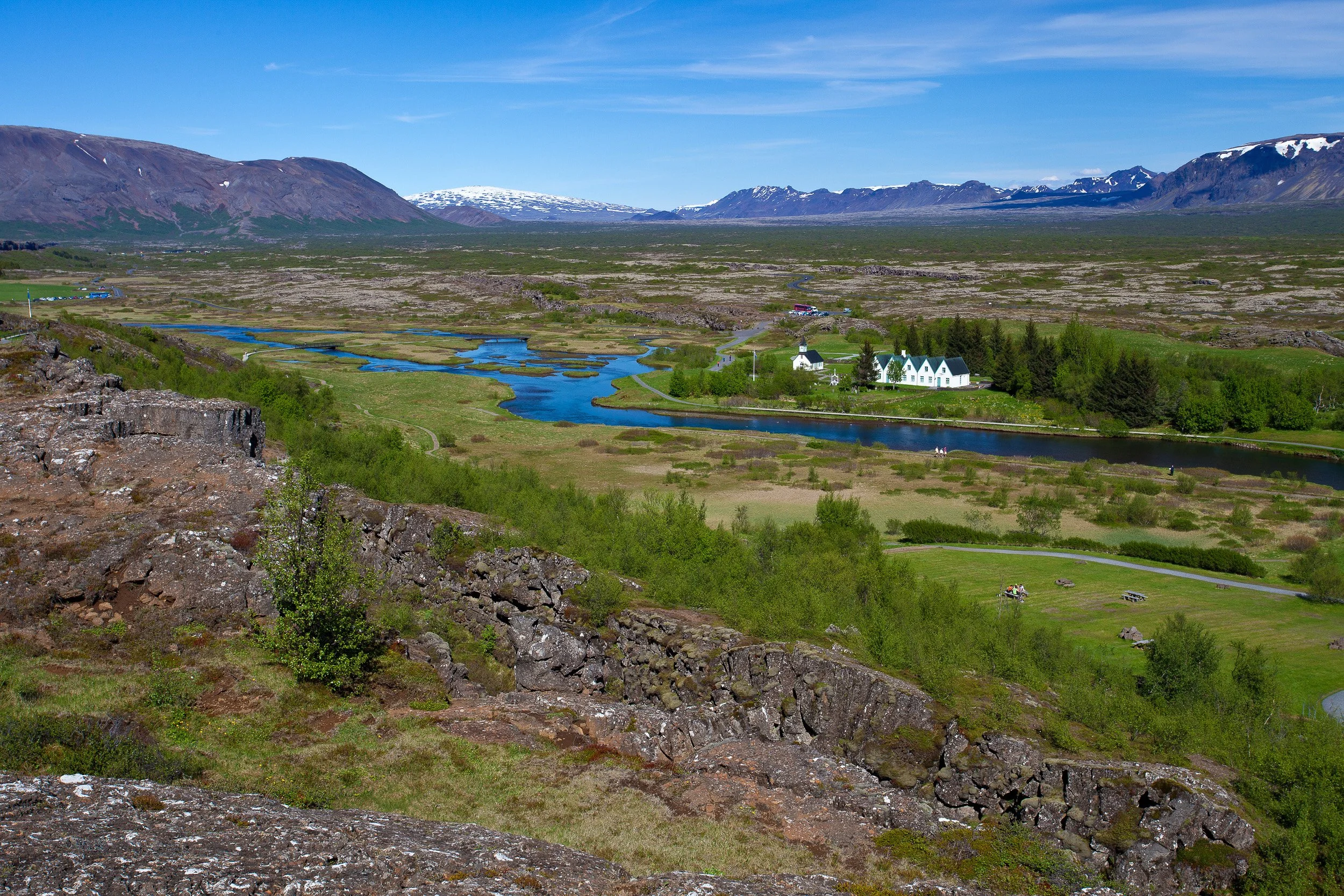 At Thingvellir National Park, the rift valley opens wide: rocky fissures in the foreground, a winding river and small church below, and snow-dusted peaks on the horizon.