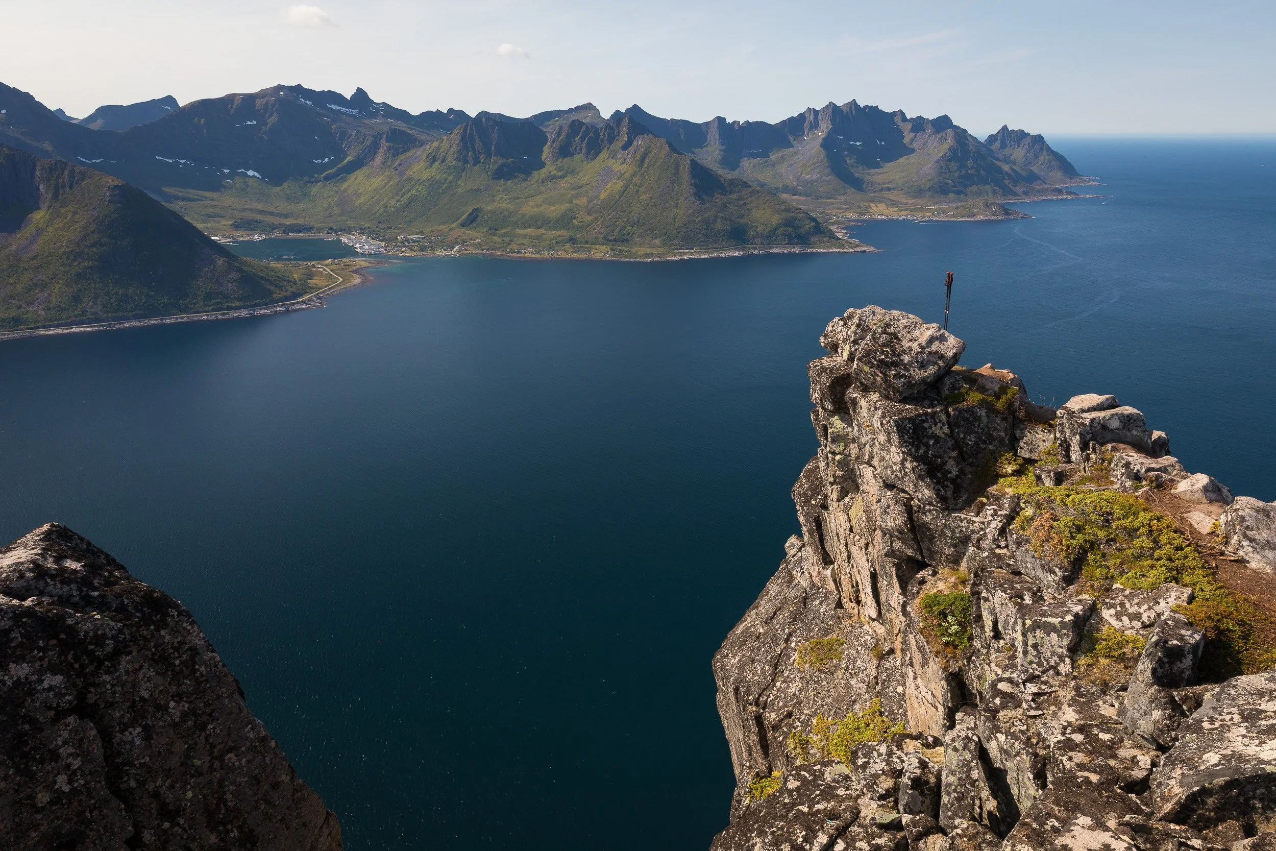 From Hesten, steep rock drops towards deep blue water and distant coastal villages, revealing the scale of Senja’s mountain-and-fjord landscape.