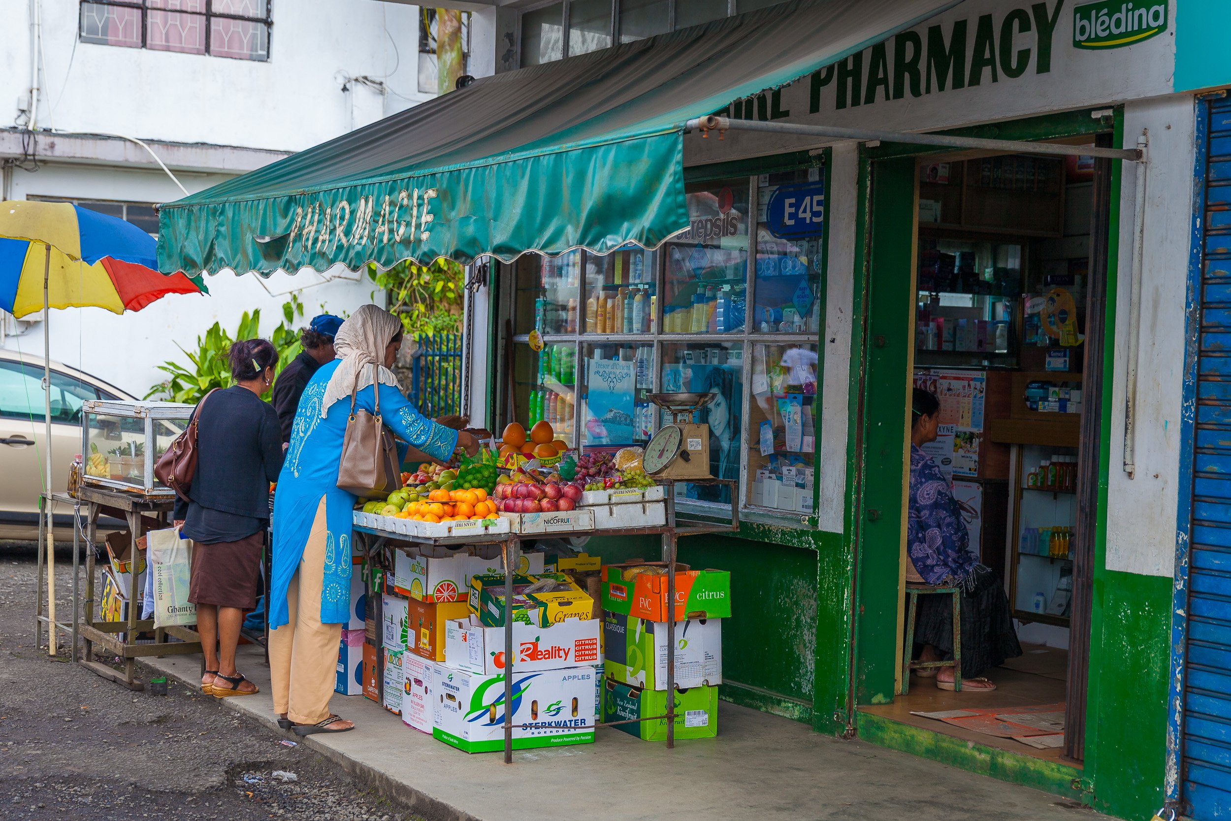 Small neighbourhood shop in Mauritius with women choosing fruit from a colourful street stall outside a shabby pharmacy.