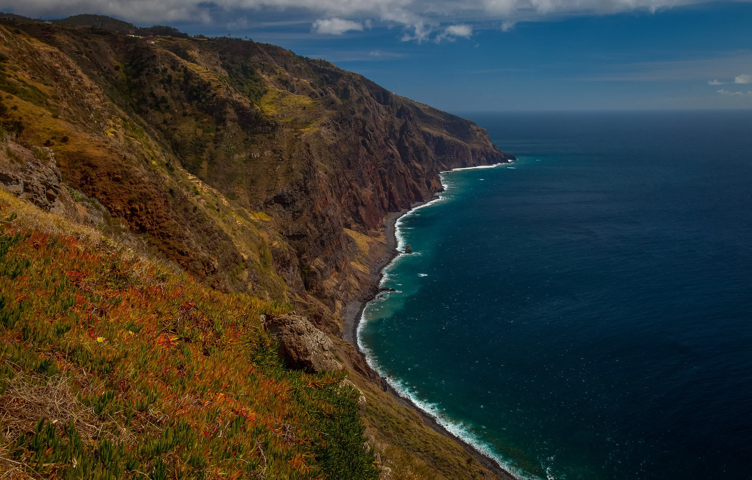 A high view along the cliffs near Ponta do Pargo, where dark Atlantic water meets a narrow shoreline beneath rugged western slopes.
