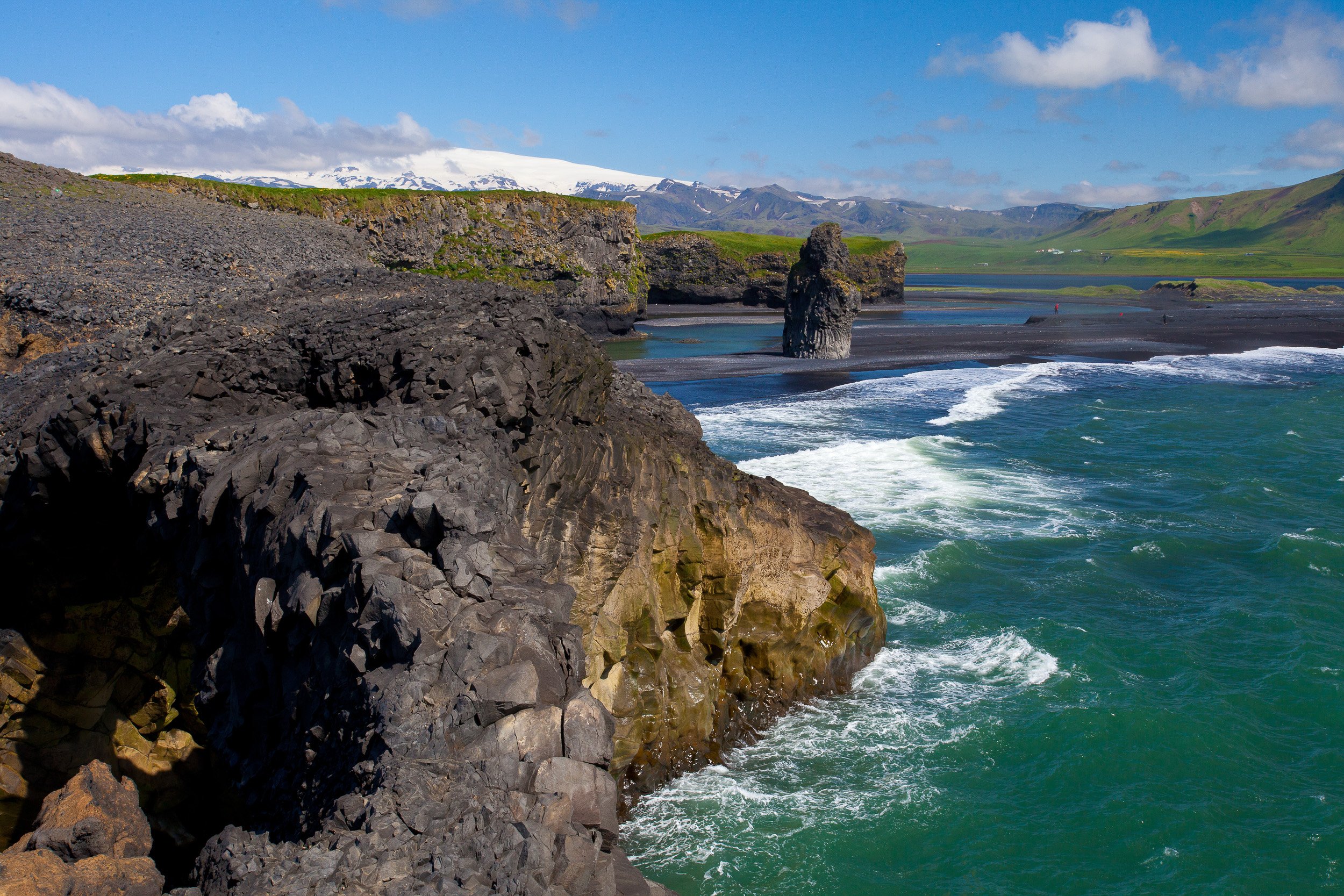 From the cliffs at Dyrholaey, dark lava rock drops to the sea as waves comb the black shoreline, sea stacks standing offshore like sentries.