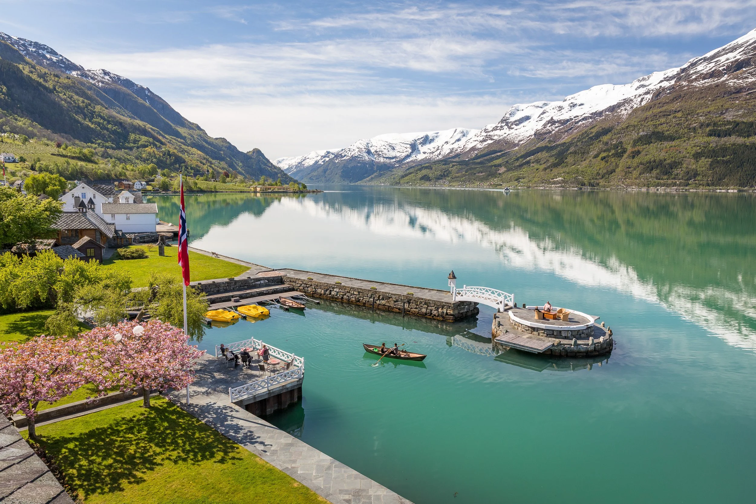 Sørfjorden near Lofthus in Hardanger — the Norwegian flag in the foreground as the fjord stretches towards snow-capped peaks, often tinted a fresh green by glacial meltwater from the Folgefonna ice cap flowing down into the surrounding waterways and 