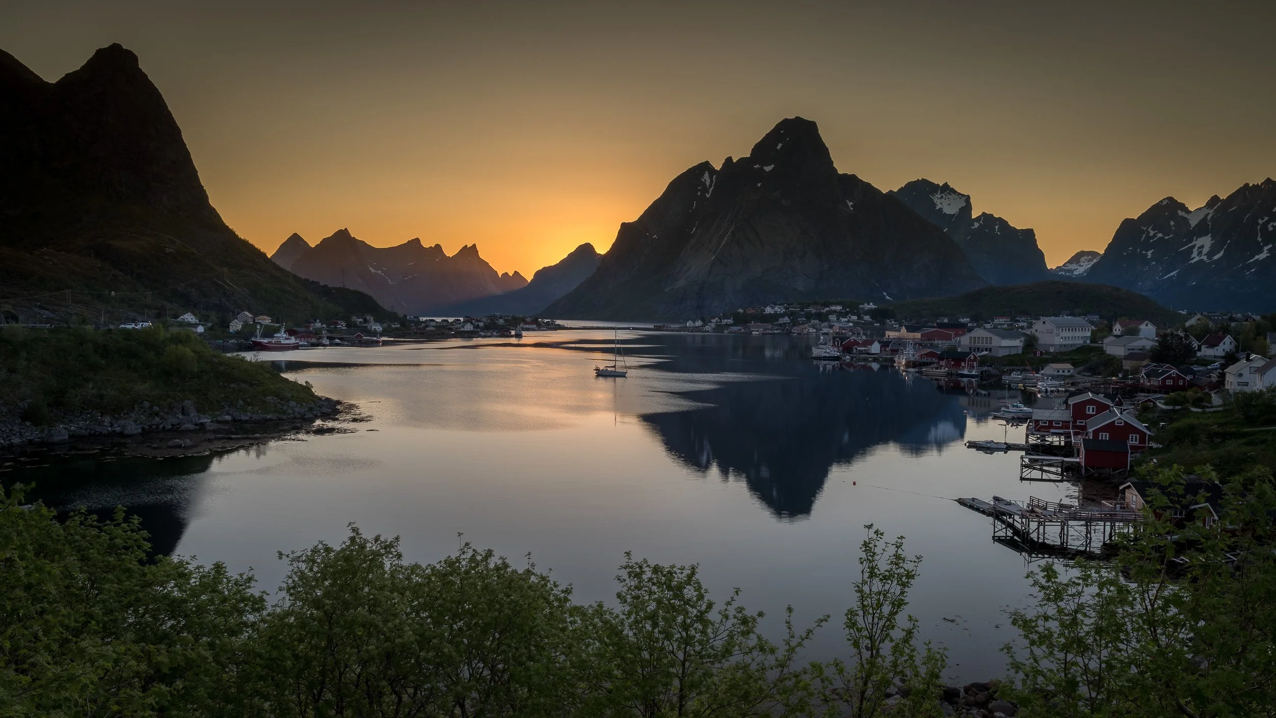 Midnight sun in Reine, Lofoten Islands, Norway – the low Arctic sun hangs above the fjord, glowing over red fishing cabins and jagged mountains. Soft golden light lingers for hours, giving an almost unreal atmosphere to this classic Lofoten fishing v