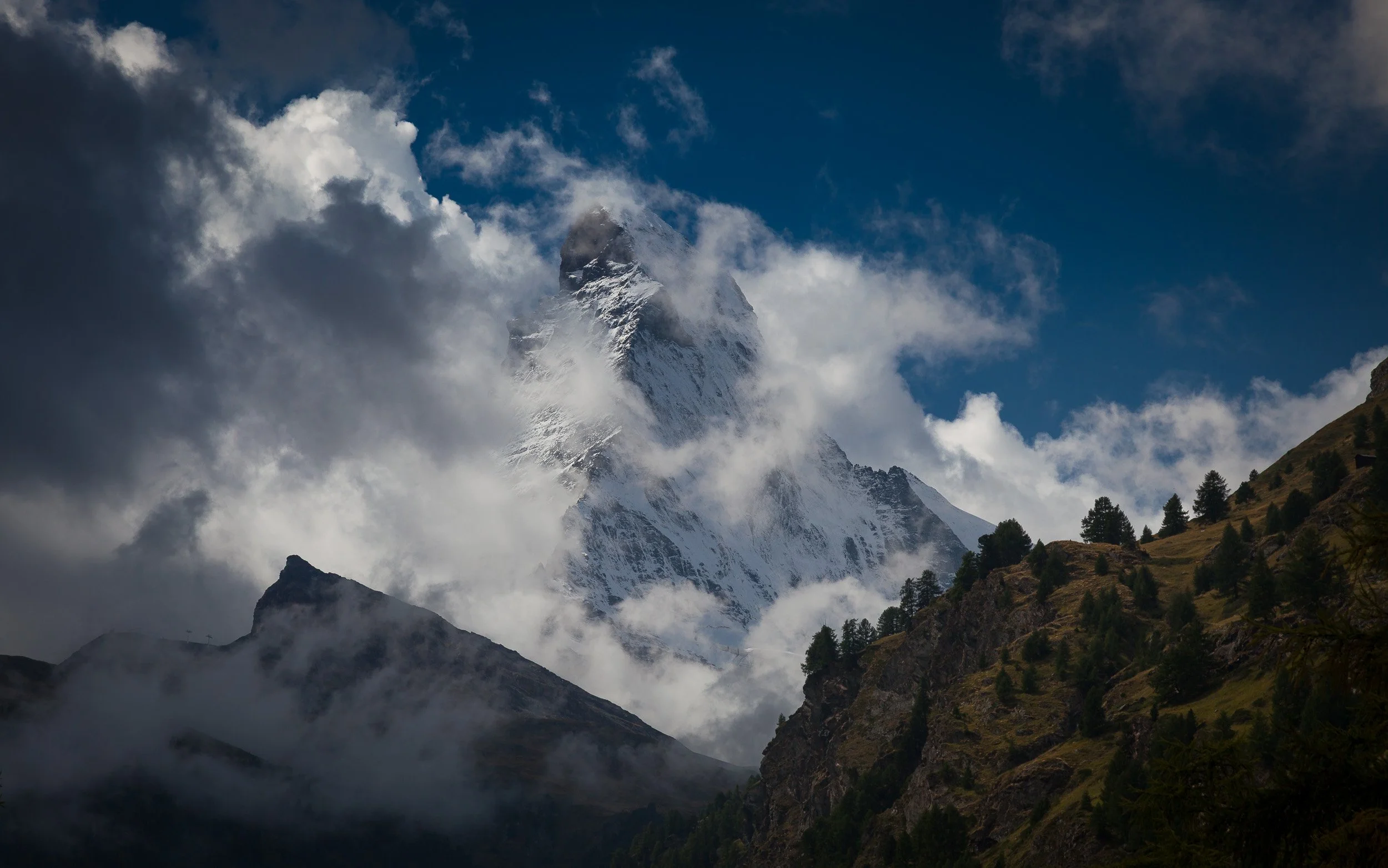 Dark storm clouds swirling around the peak of the Matterhorn above shadowed forests on the lower slopes.