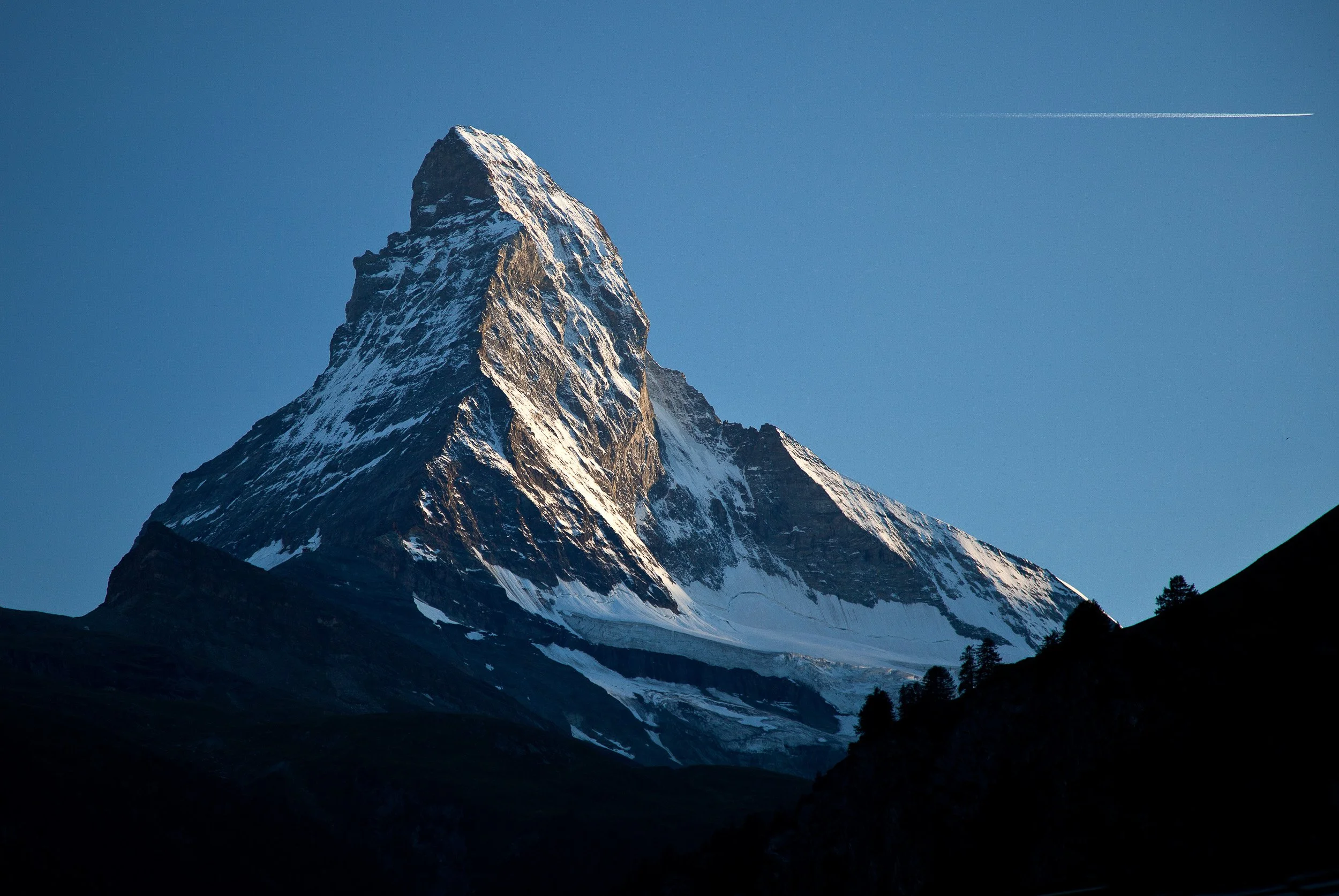 Sharp, snow-dusted profile of the Matterhorn lit by low evening sun, with a single jet trail crossing the clear alpine sky.