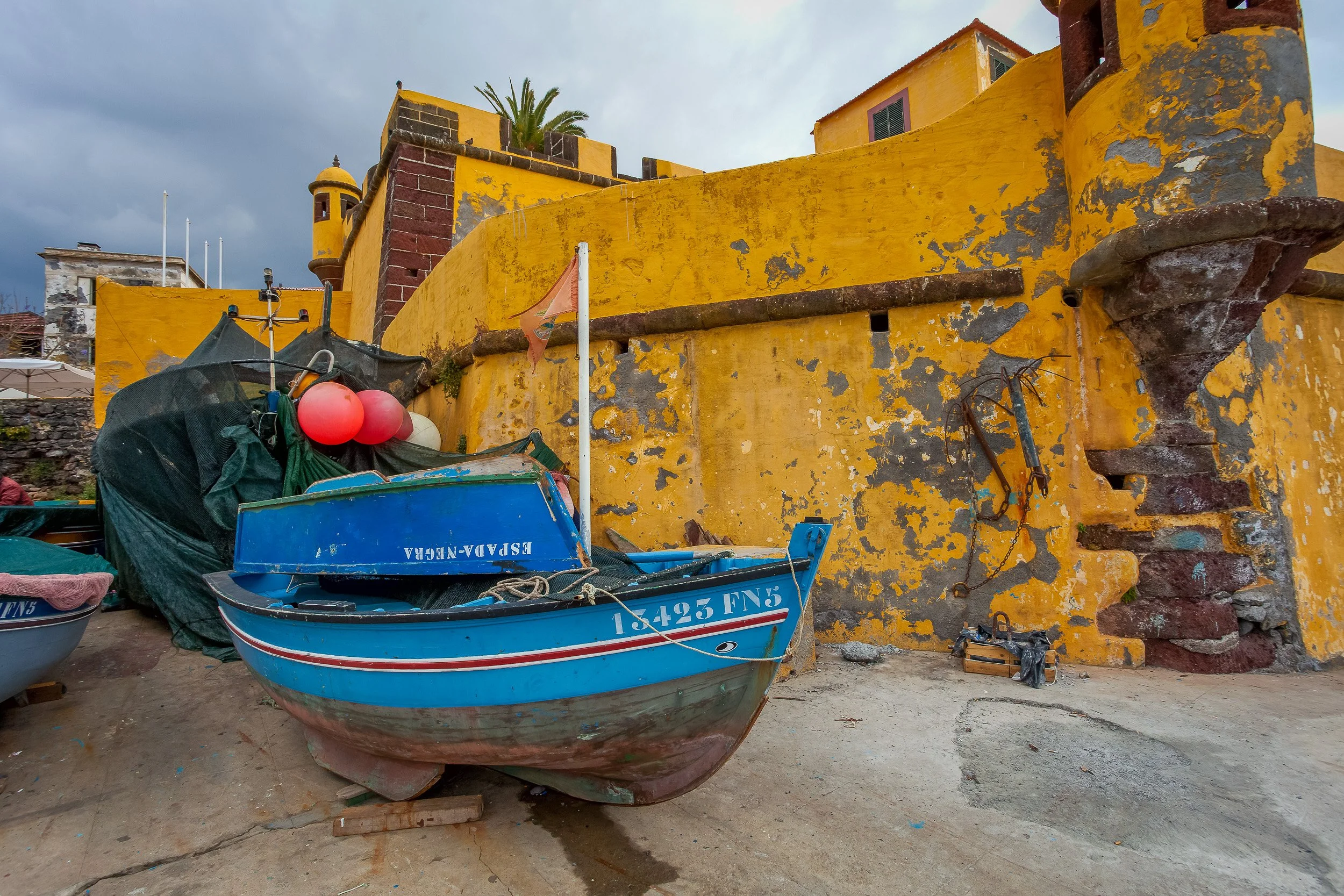 A blue fishing boat rests below the weathered yellow walls of Forte de São Tiago in Funchal, with peeling paint, nets and floats adding texture to the harbour scene.