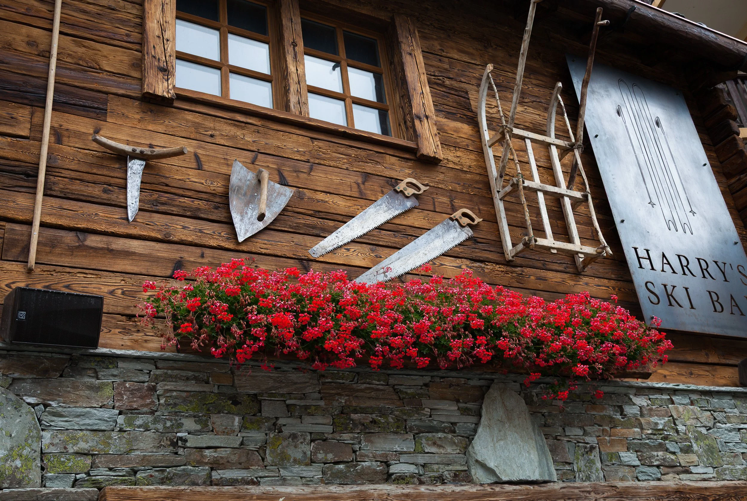Close-up of a chalet façade decorated with old wooden tools and a cascade of red flowers spilling over a stone wall.