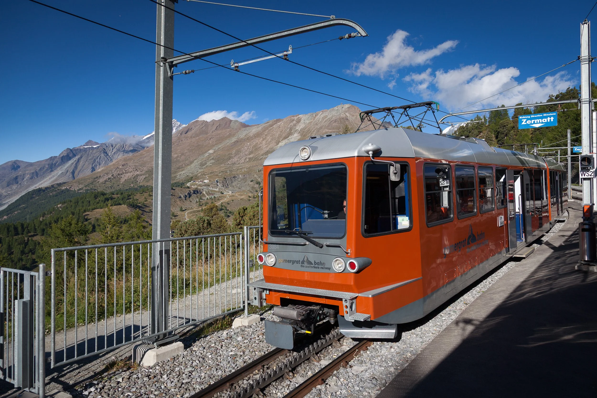 Orange Gornergrat cogwheel train standing at a mountainside station above Zermatt under a clear blue sky.
