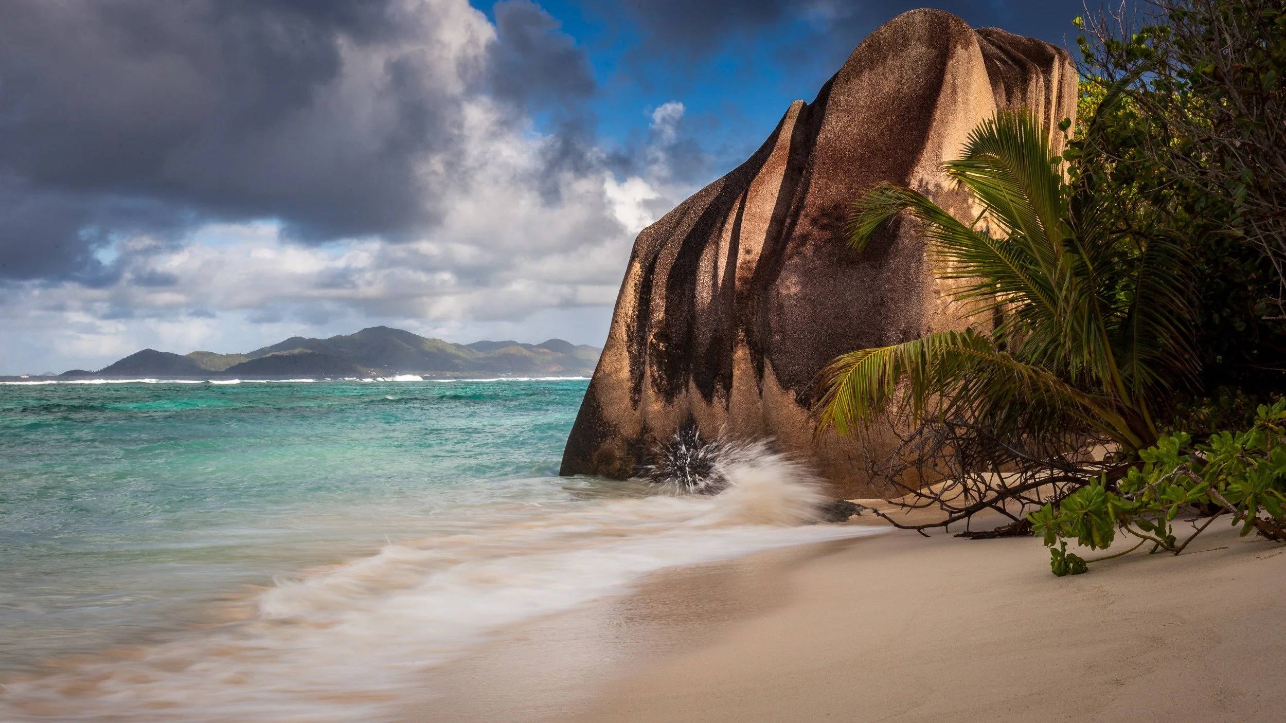 Warm sunlight hitting a large granite boulder at Anse Source d’Argent, with pale sand, turquoise water, tropical vegetation and dark rain clouds over distant islands.
